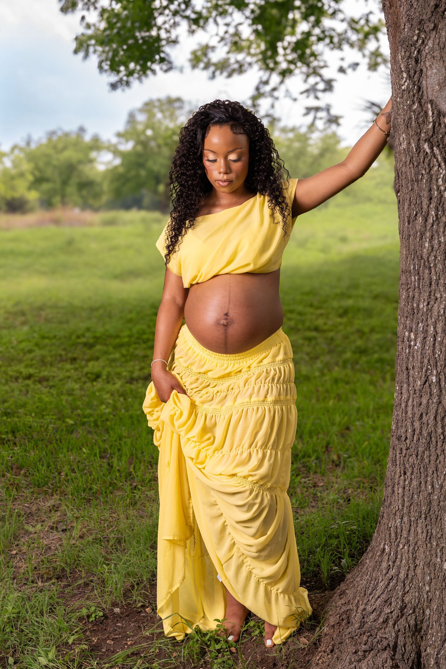 Pregnant Black woman in yellow outfit leaning against tree in green field, showing baby bump during outdoor maternity photoshoot.