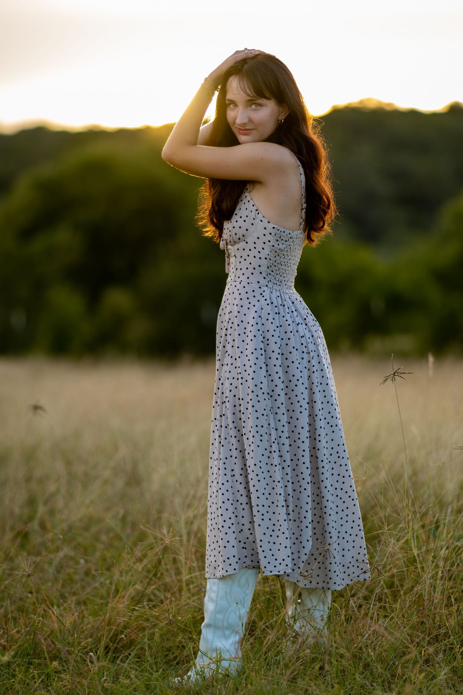 Young woman in polka dot sundress and white boots stands in golden field at sunset, glancing back over shoulder.