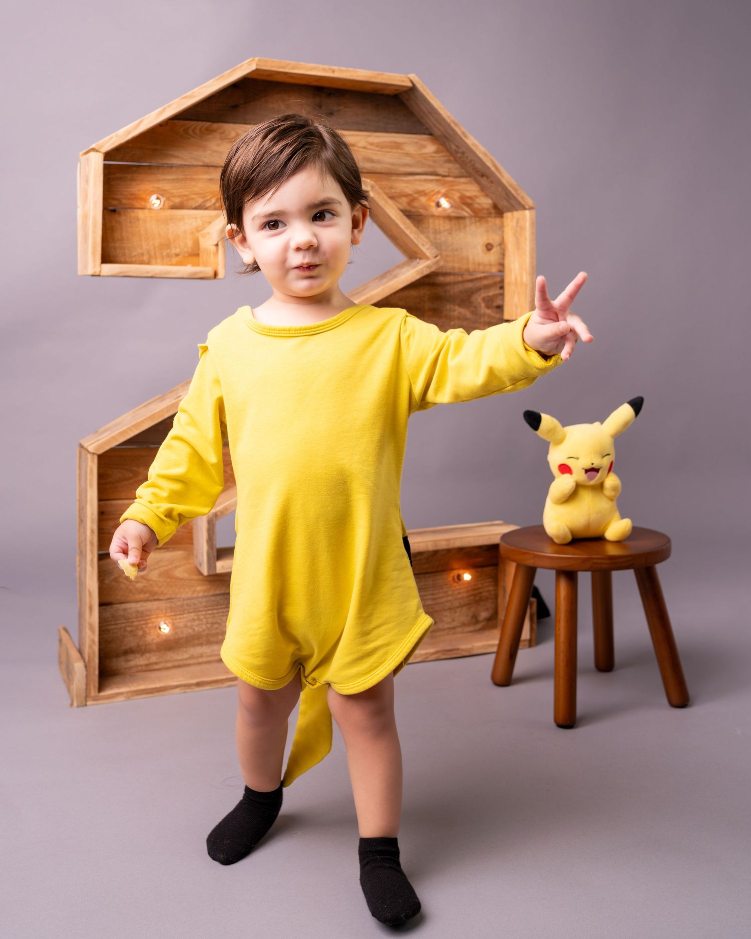 Young child in yellow romper poses next to a Pikachu plush toy on a wooden stool with decorative shelf backdrop.