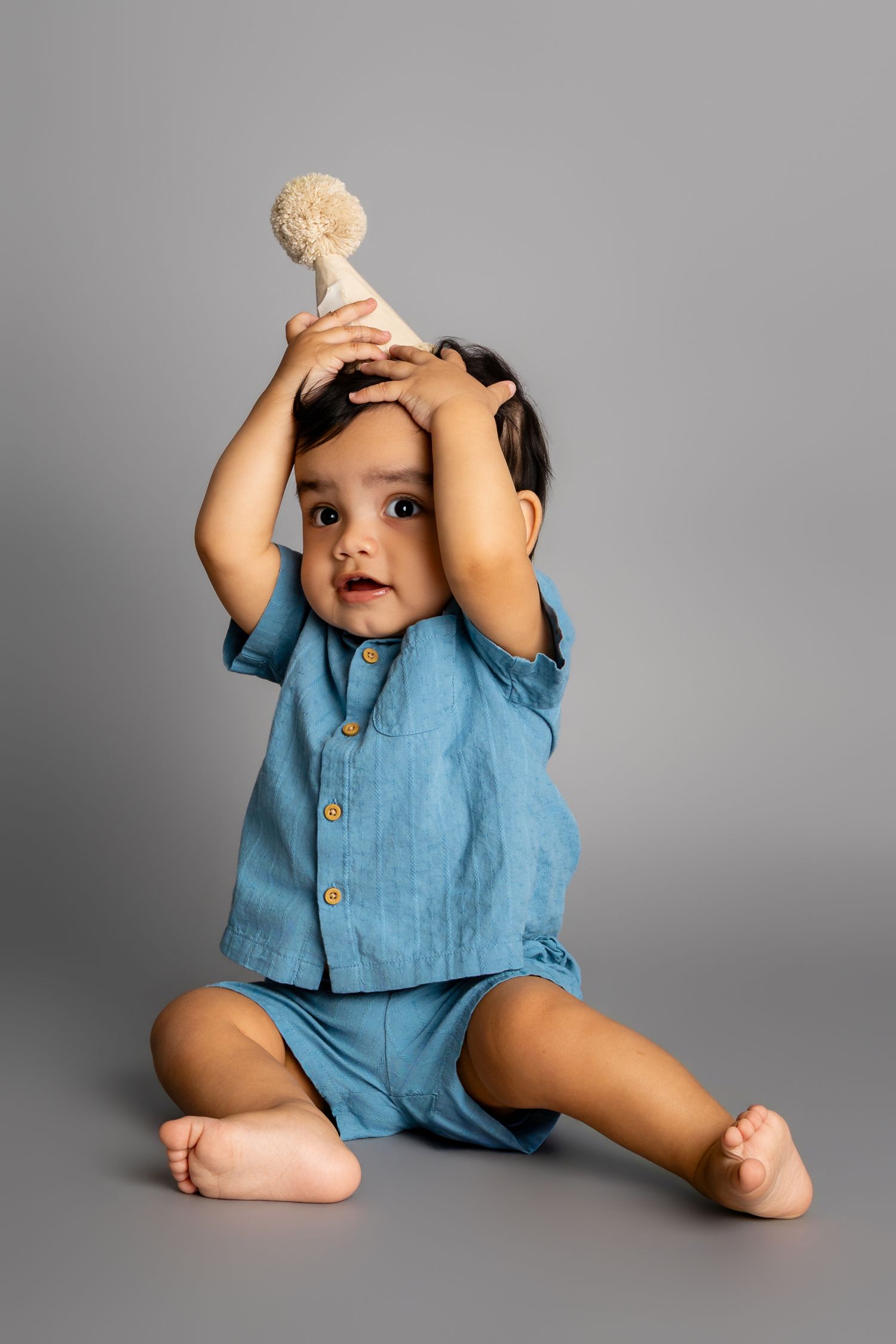 Adorable baby in blue linen outfit sitting on gray background, wearing a cream pom-pom party hat.