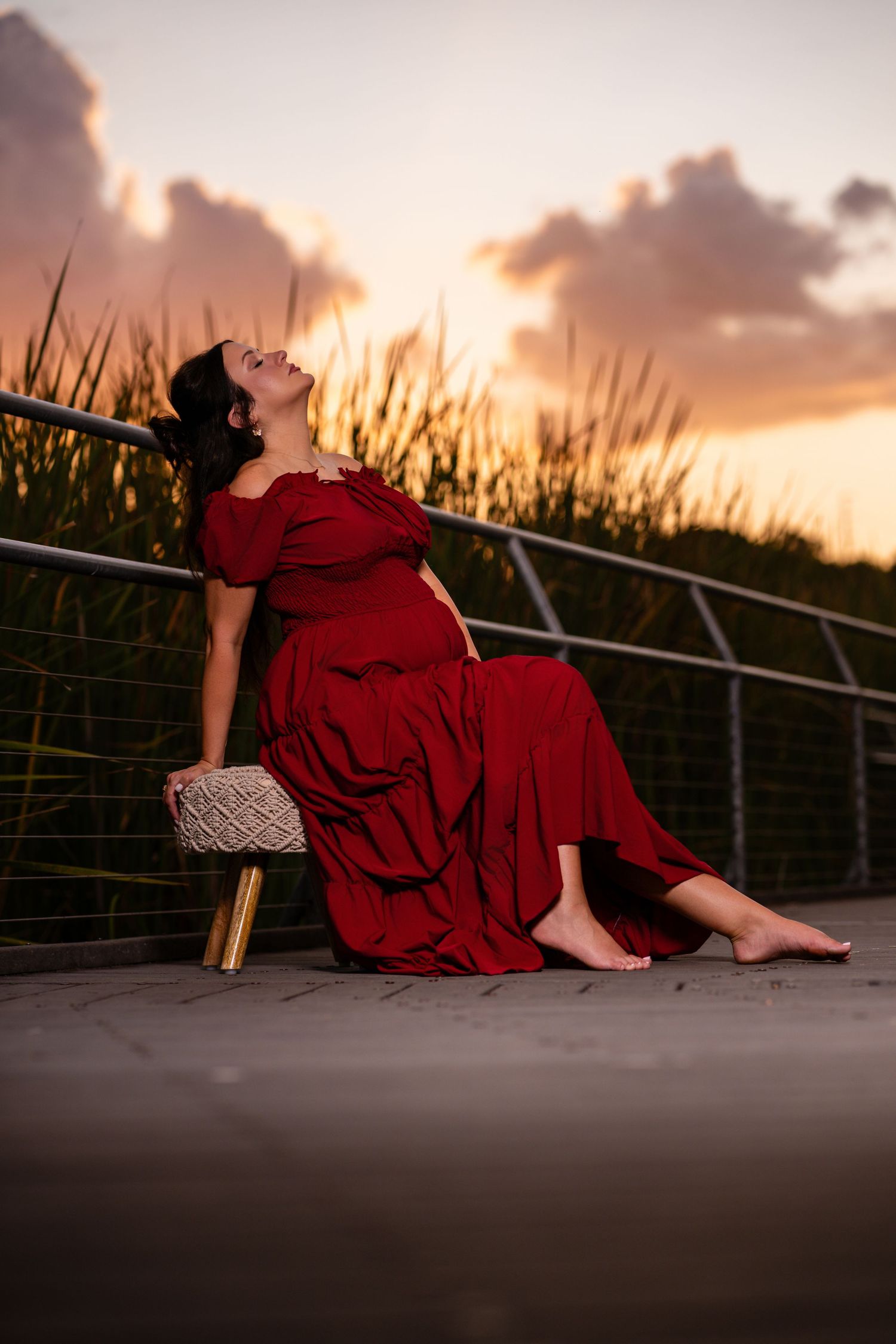 Pregnant woman in red gown sitting barefoot at sunset, leaning back gracefully with tall grass and colorful sky behind her.