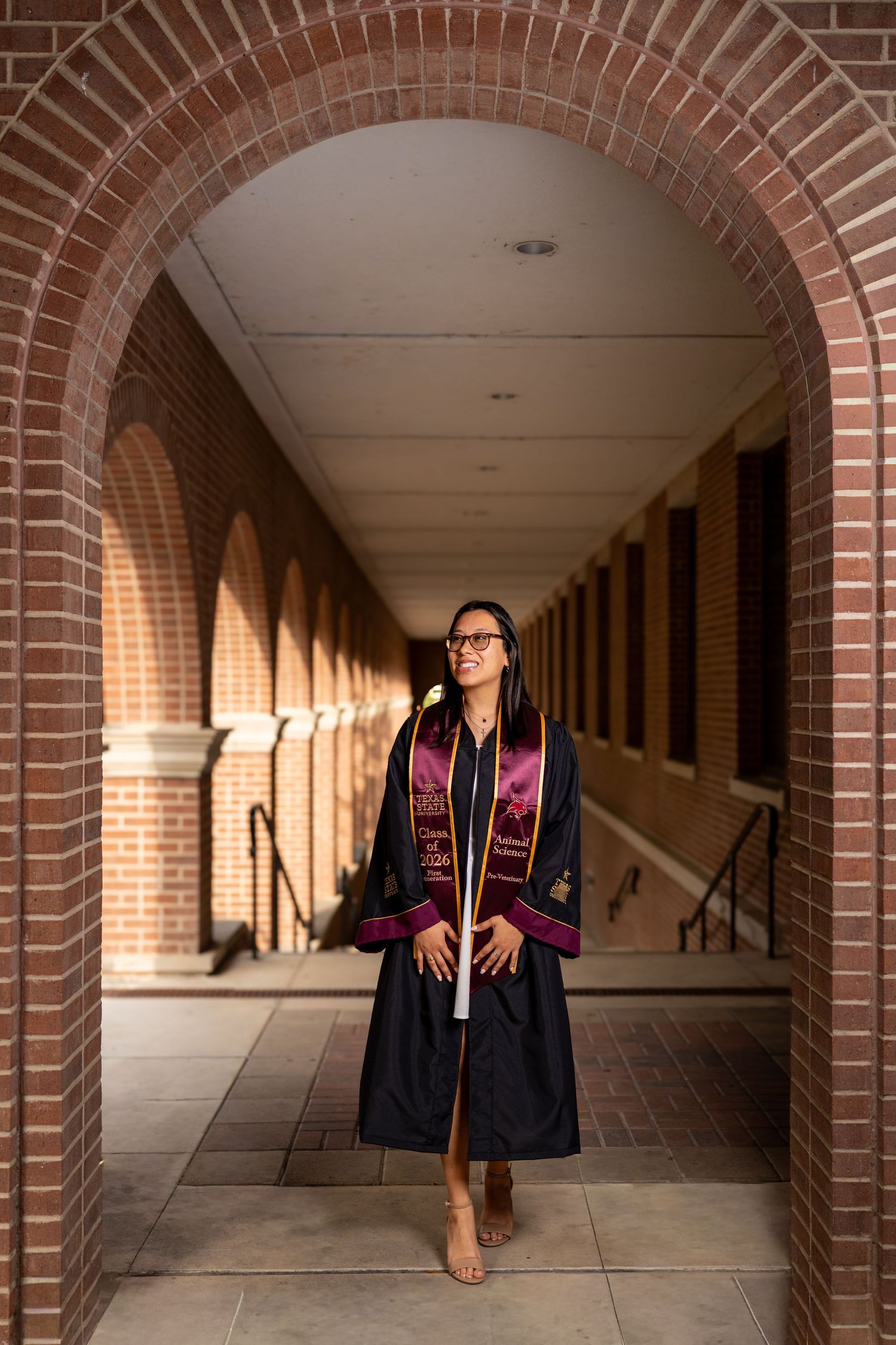 Graduate in black gown and maroon stole poses under brick archway at university building.