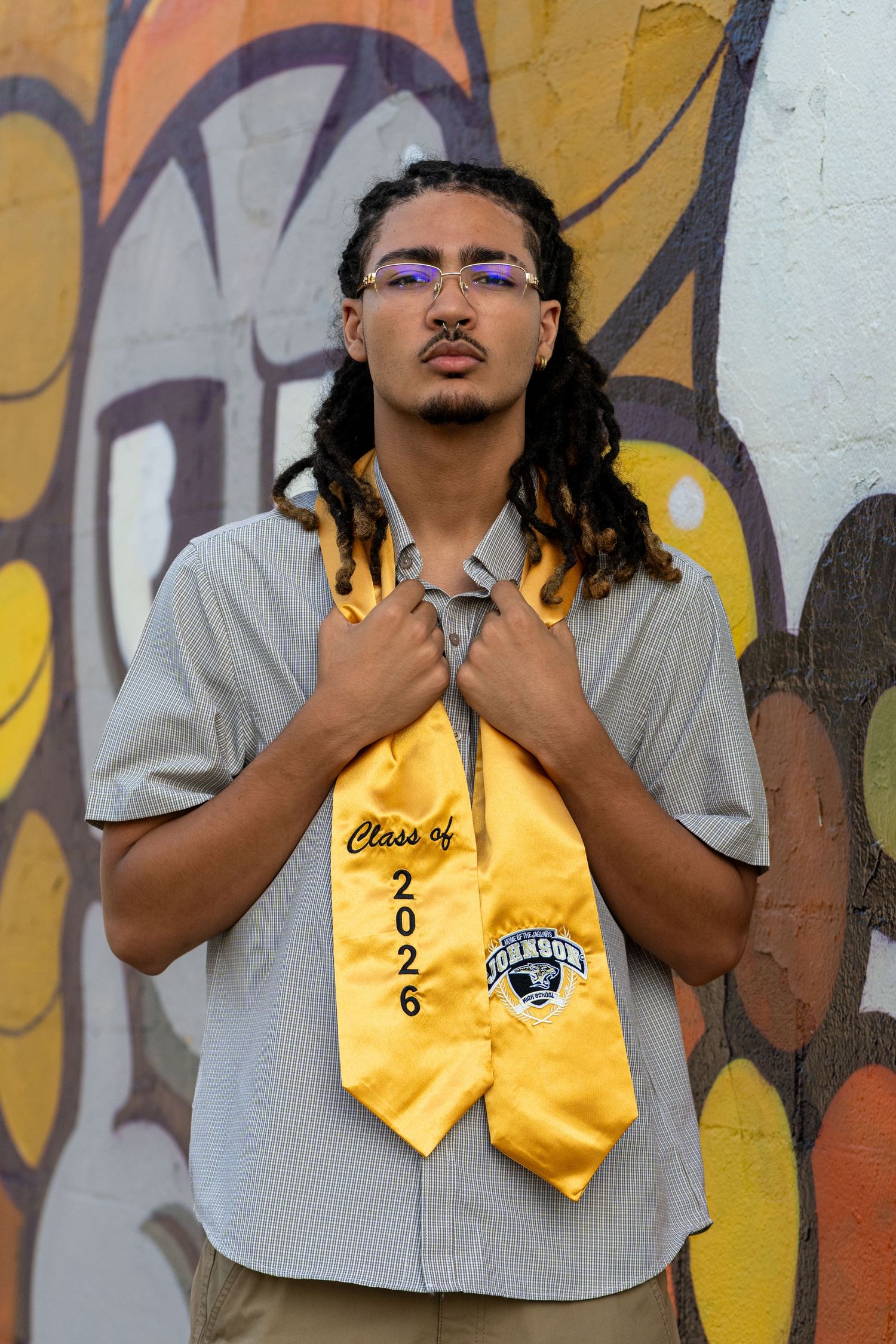 Young man wearing Class of 2026 gold graduation stole poses confidently in front of colorful graffiti wall.