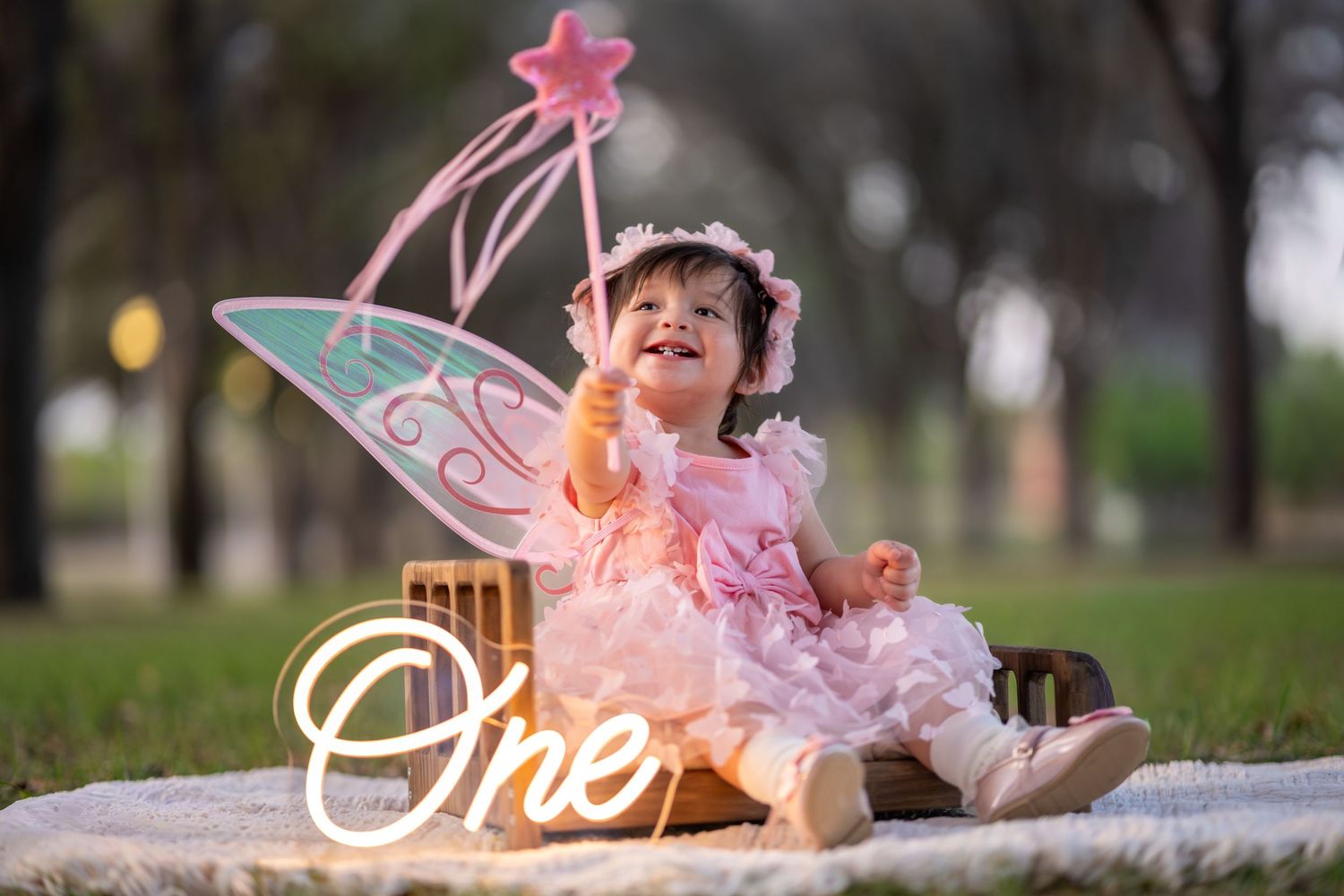 Smiling baby girl in pink fairy costume with wings and wand celebrates first birthday outdoors.