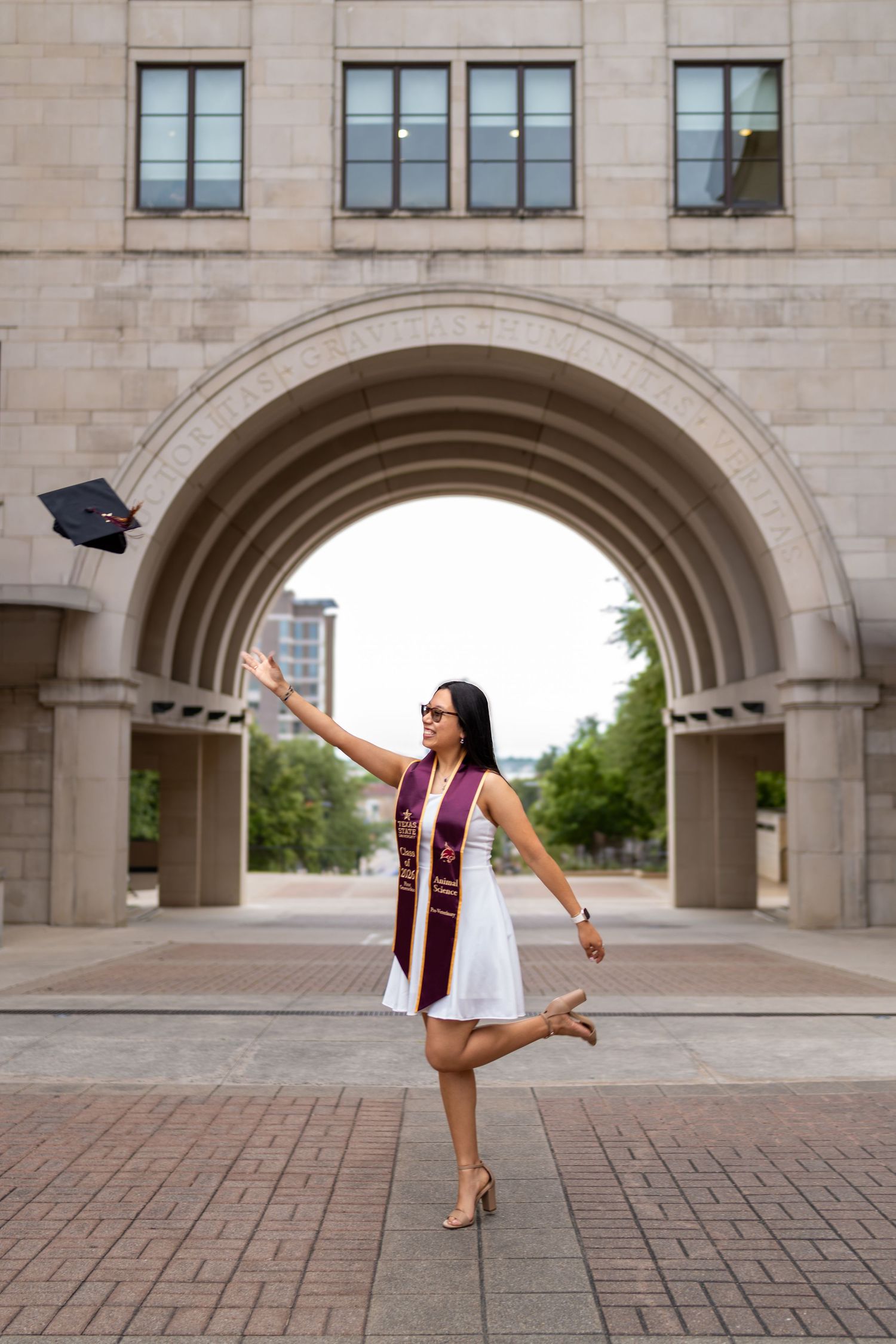 Graduate in white dress tosses cap under stone arch, celebrating commencement in maroon honor stole.
