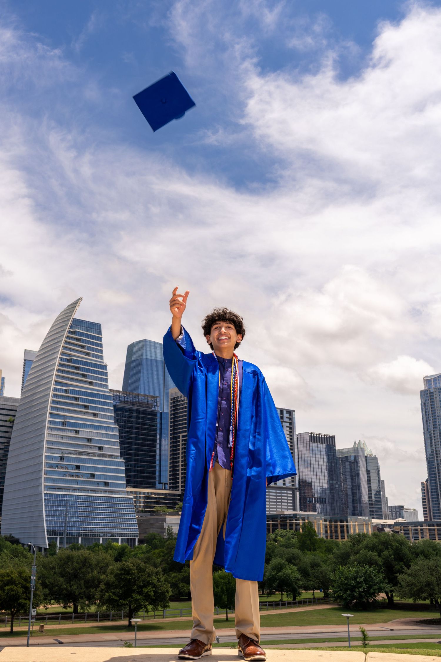 Graduate in blue cap and gown tosses mortarboard into sky with city skyline backdrop on sunny day.