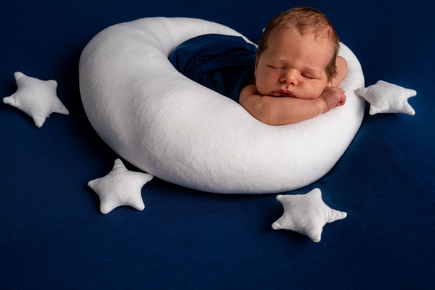 Newborn baby sleeping peacefully on a white crescent moon pillow surrounded by star cushions on a dark blue background.