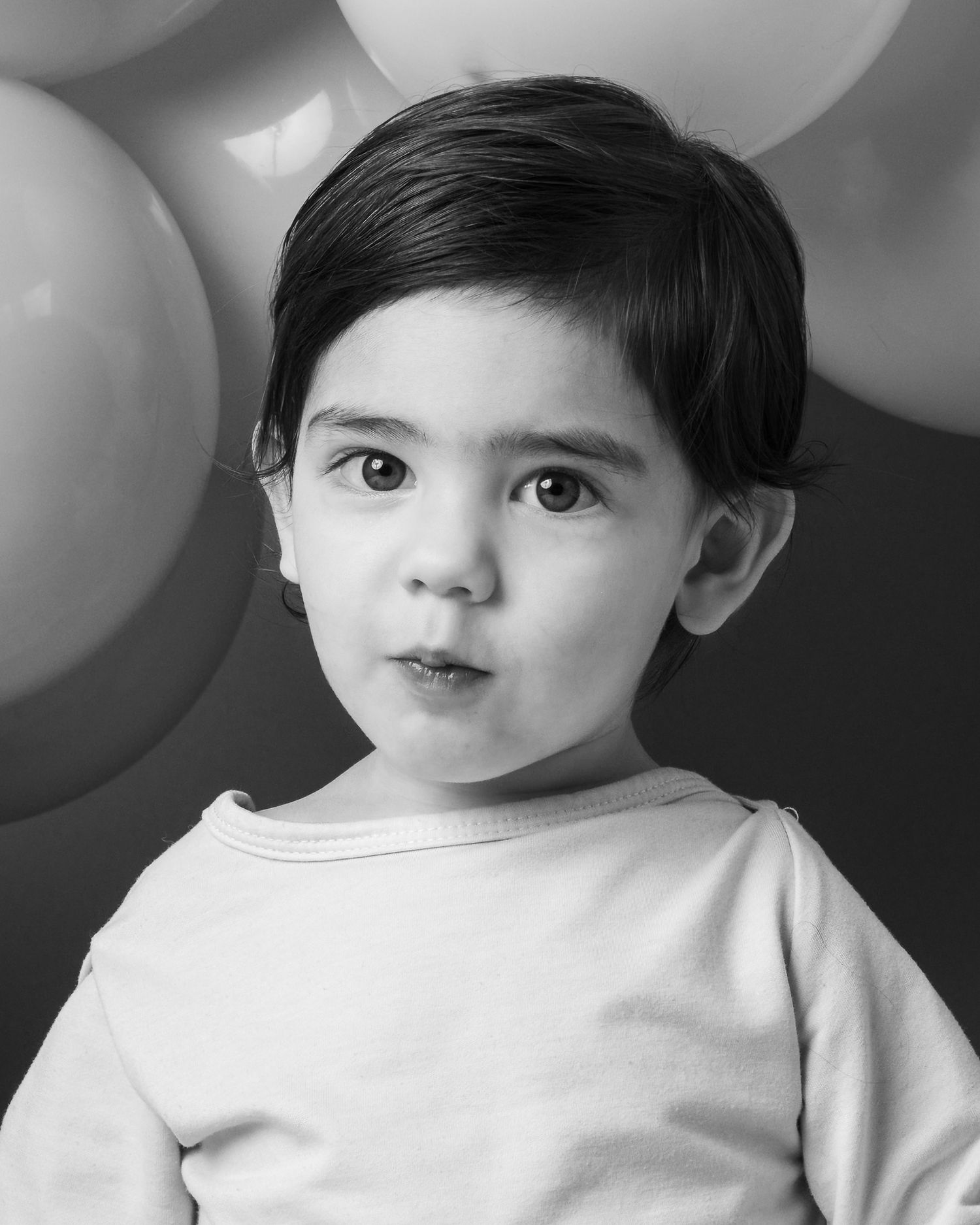 Black and white portrait of a young child in a white shirt against a blurred balloon background.
