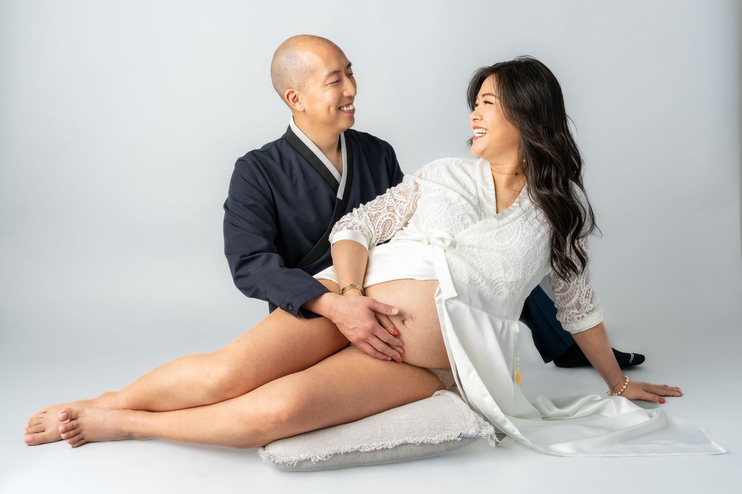 Couple in a maternity photoshoot, man in navy robe holding pregnant woman's belly, sitting on white backdrop.