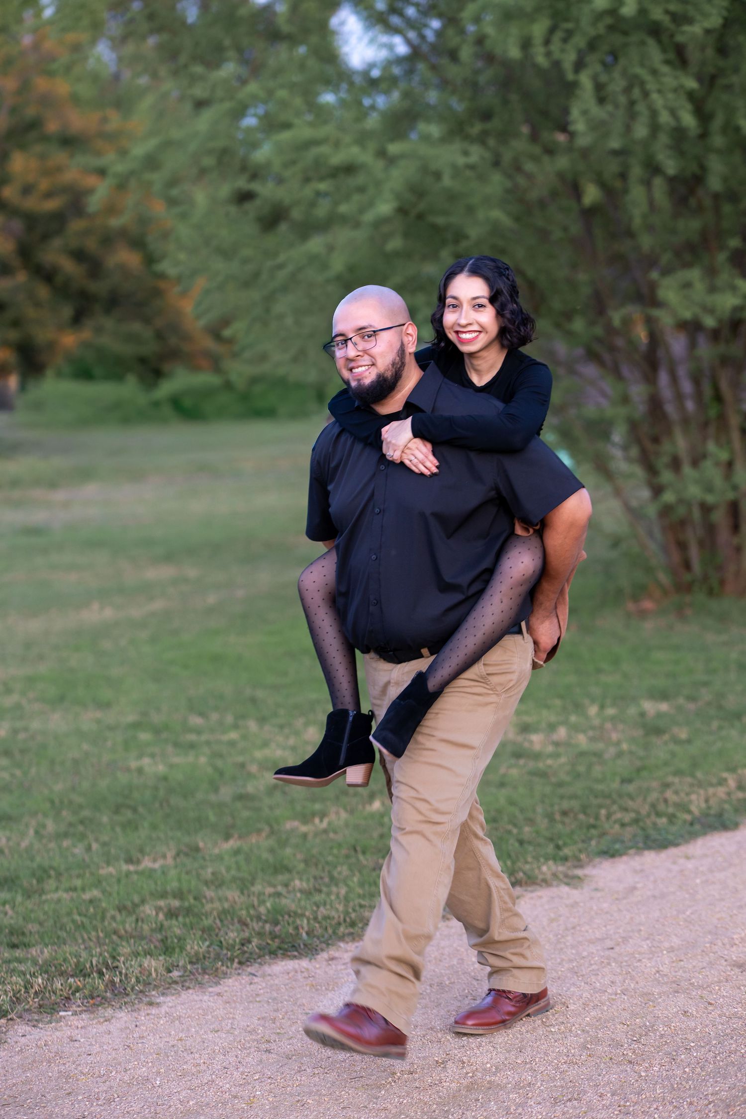 A playful couple shares a piggyback ride moment on a dirt path surrounded by green trees and grass.