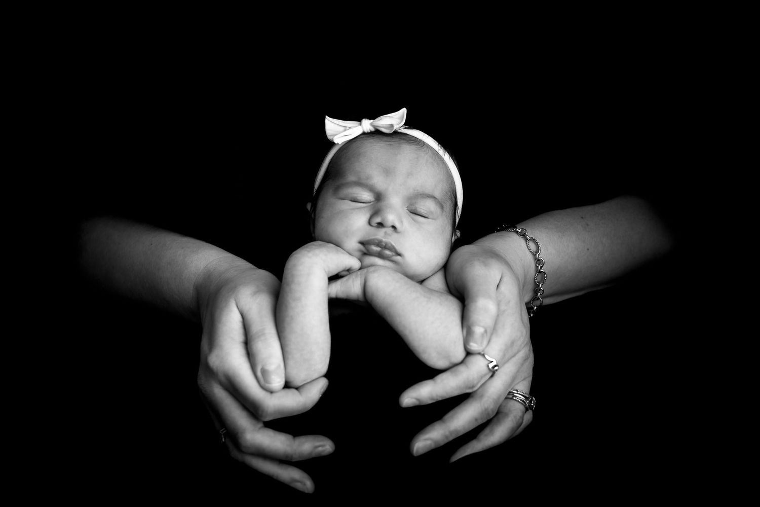 A sleeping newborn baby rests peacefully in black and white while being gently cradled by adult hands.