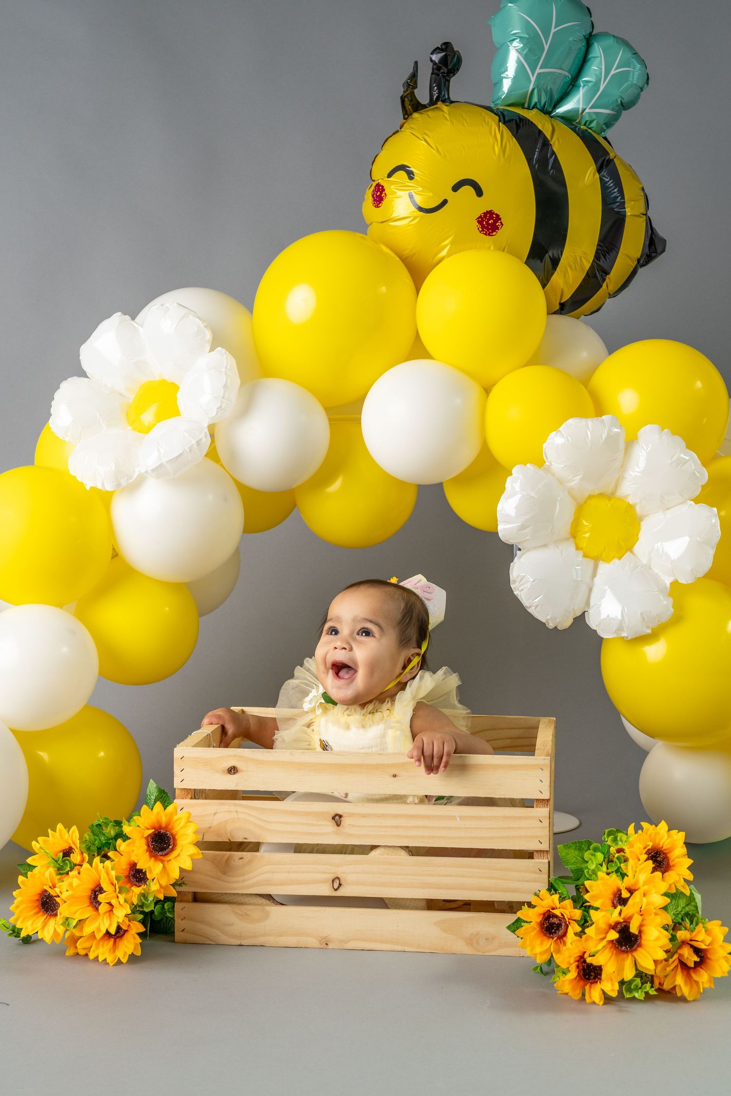 Happy baby in wooden crate surrounded by yellow and white balloon arch with bee balloon and sunflowers.