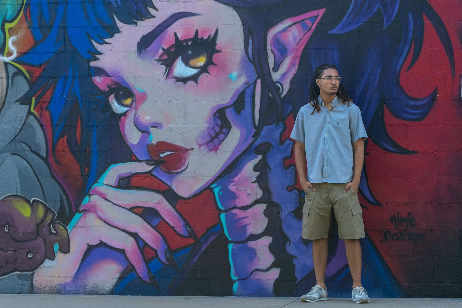 A young man stands in front of a vibrant graffiti mural featuring an anime-style girl with elf ears on a colorful wall.