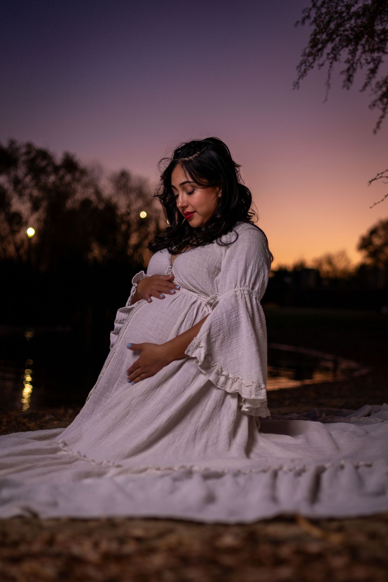 A woman in a flowing white dress sits on the ground during sunset in a desert landscape.