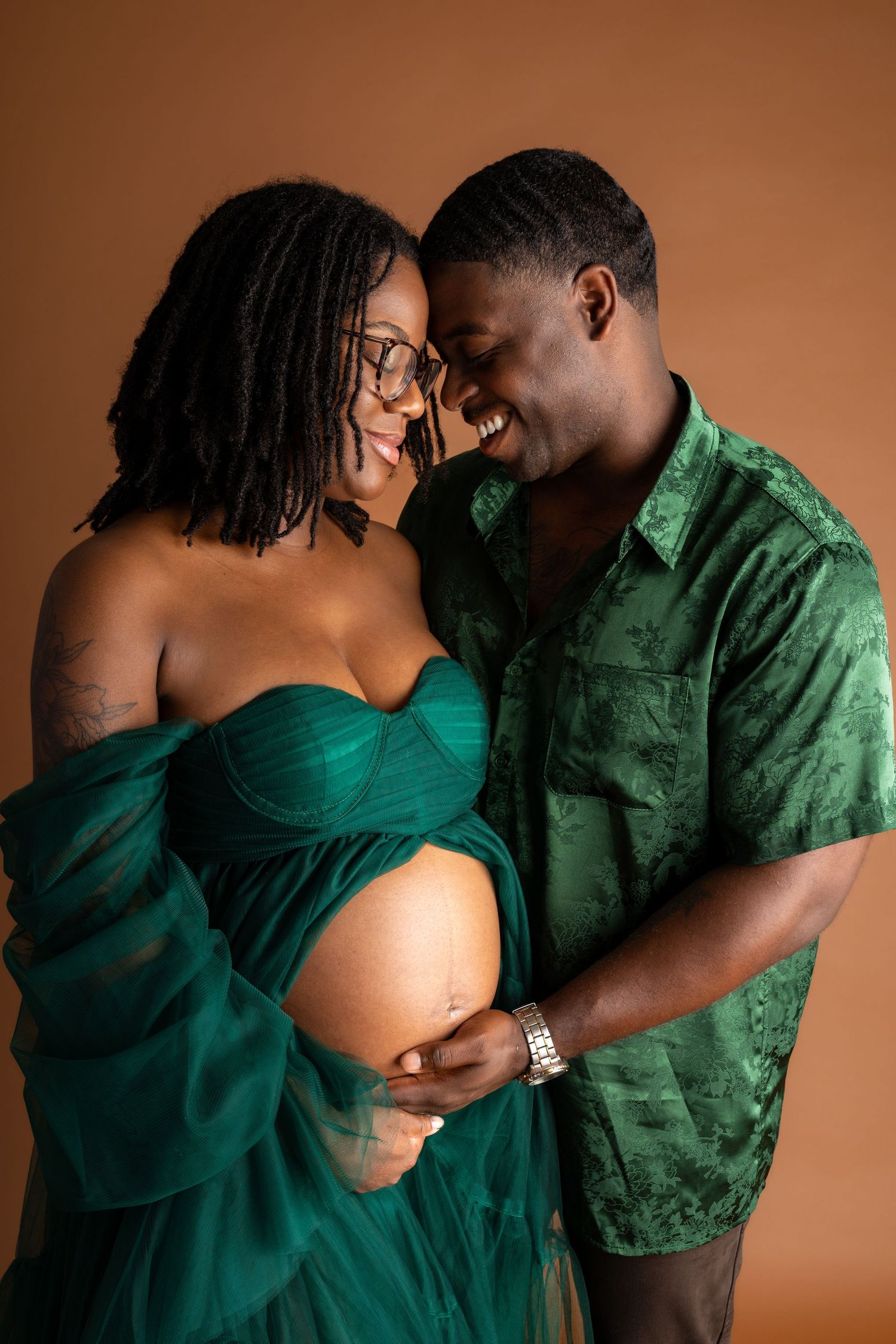A joyful pregnant couple in matching green outfits share an intimate moment, smiling together against a warm brown background.