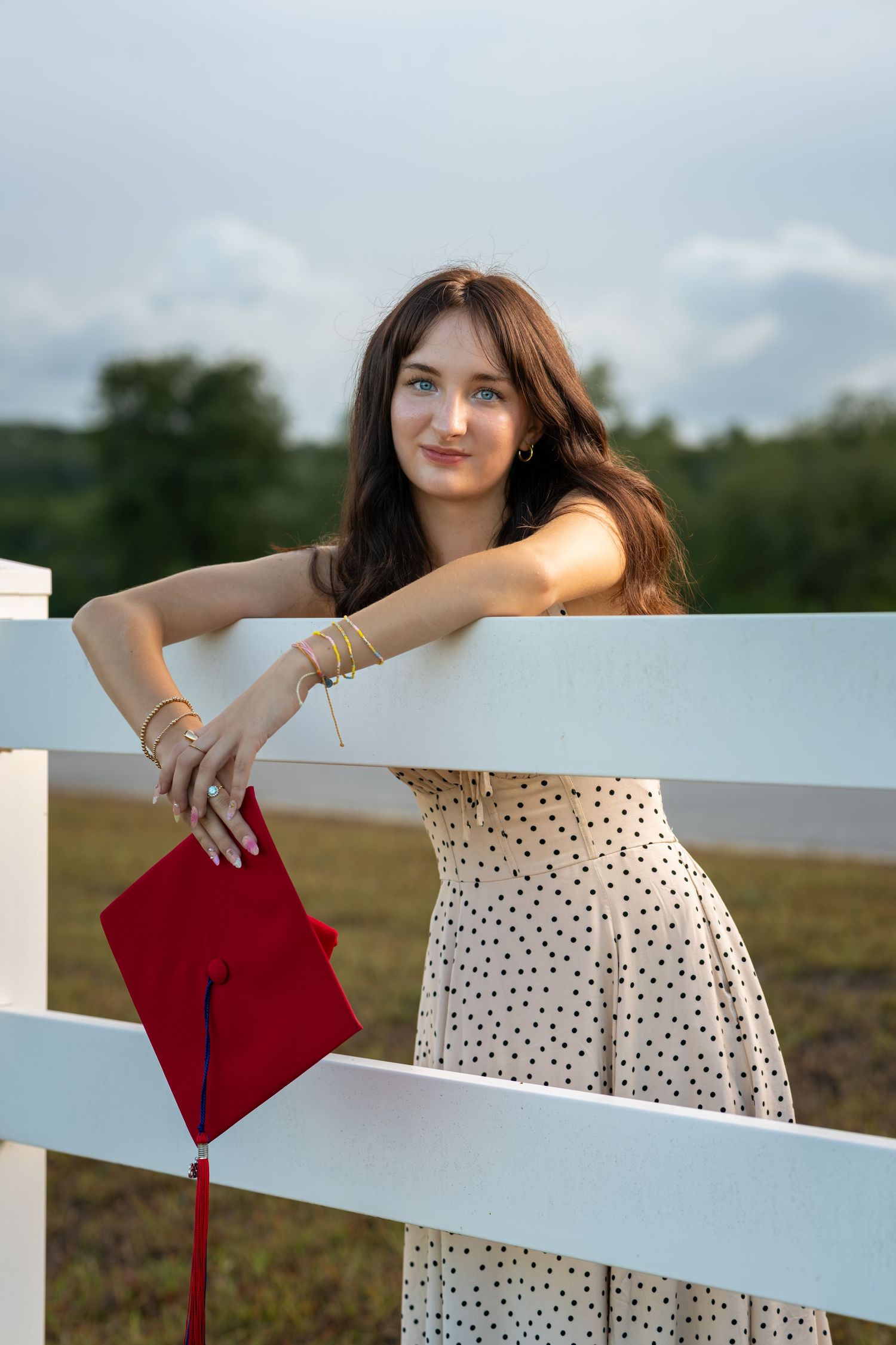 Young woman in polka dot dress holding red graduation cap, leaning on white fence outdoors.