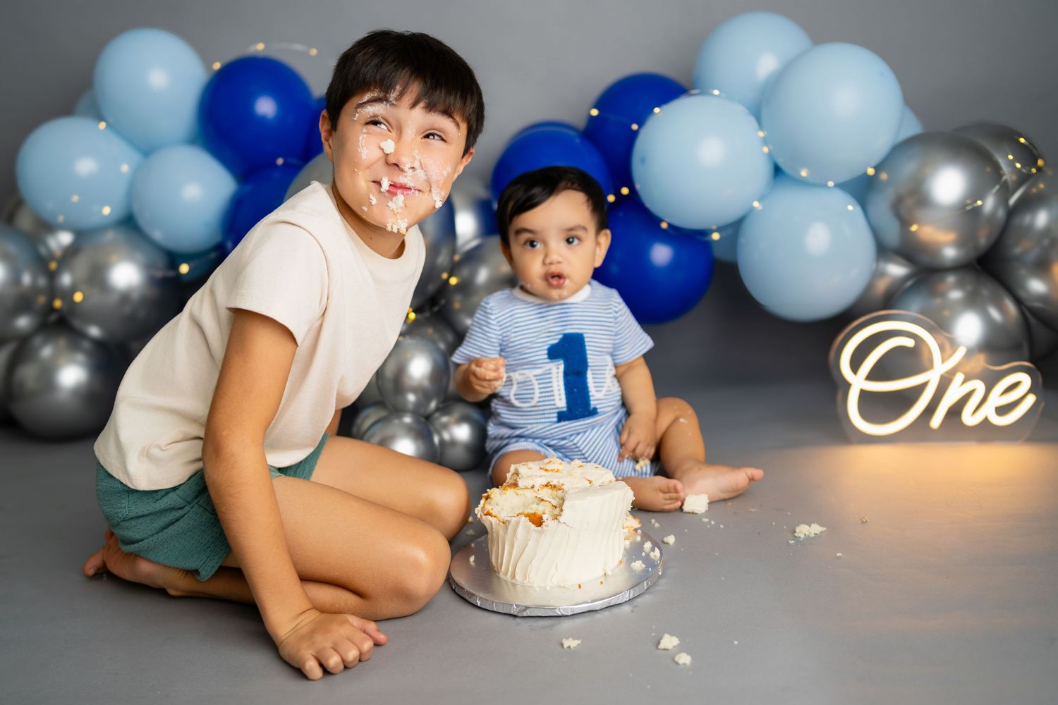 Two boys celebrate a first birthday cake smash with blue balloons and an One neon sign in the background.
