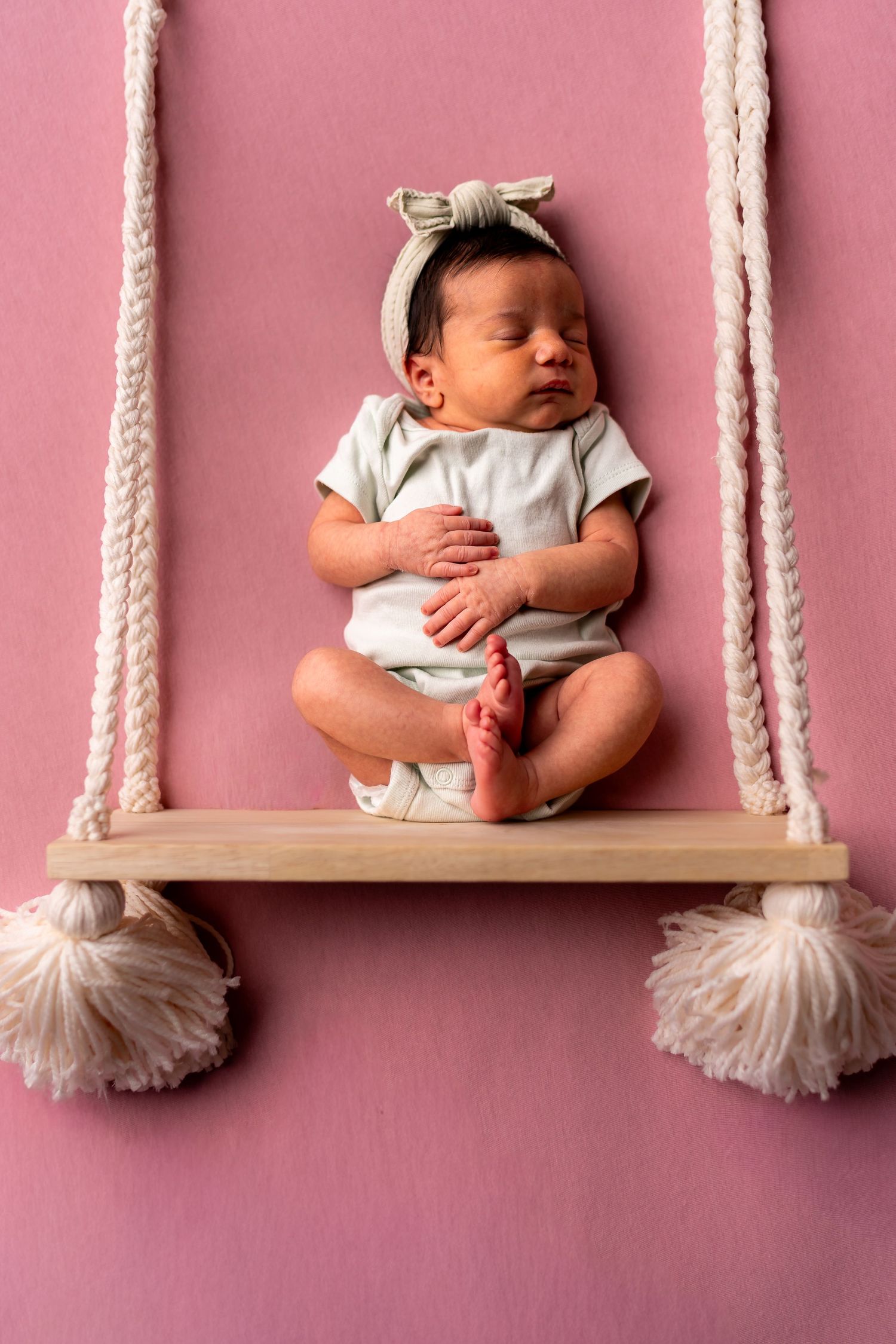 Baby resting on wooden swing with macrame ropes against pink background.