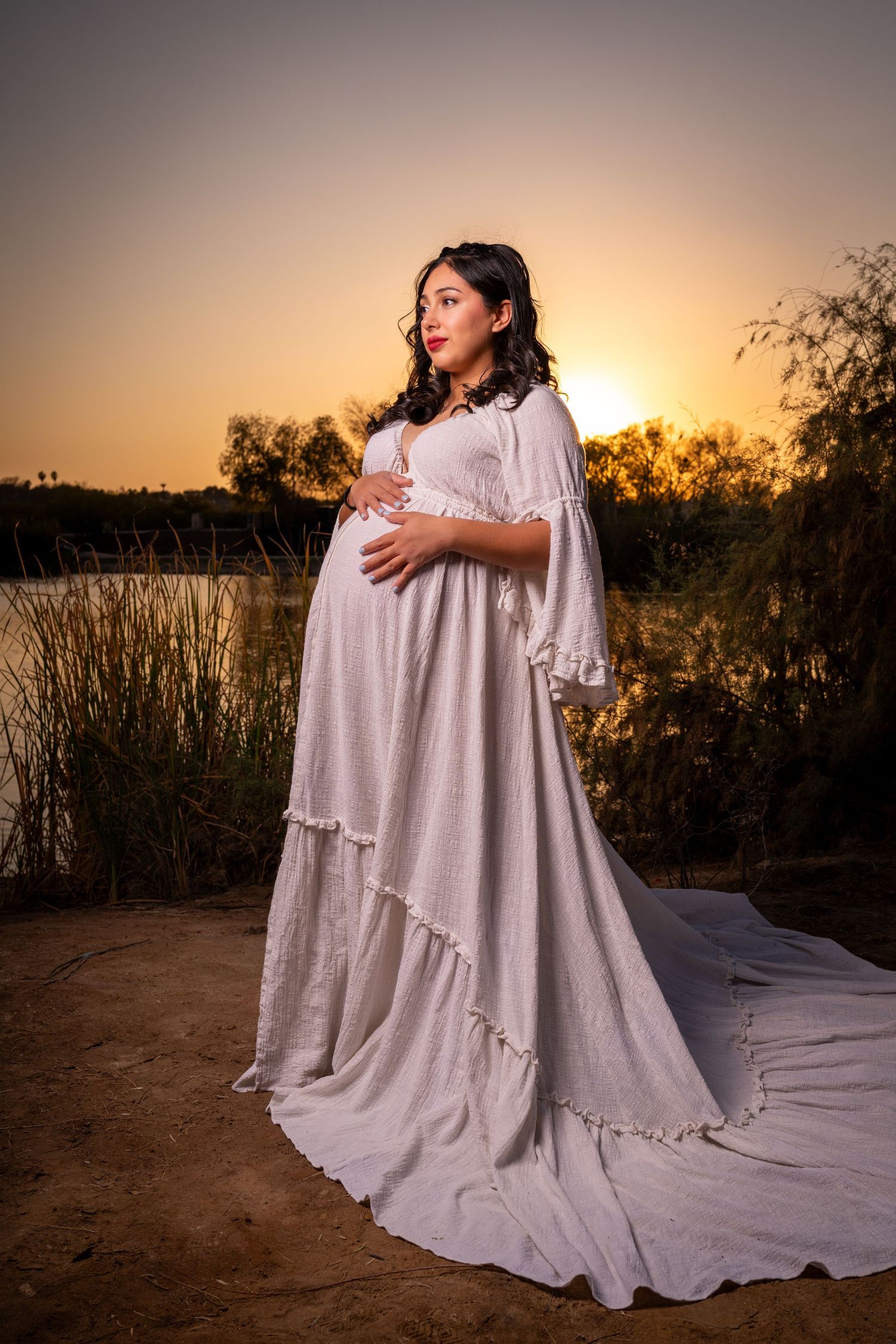 A maternity photo session at sunset shows a woman in a flowing white dress against a golden sky and natural landscape.