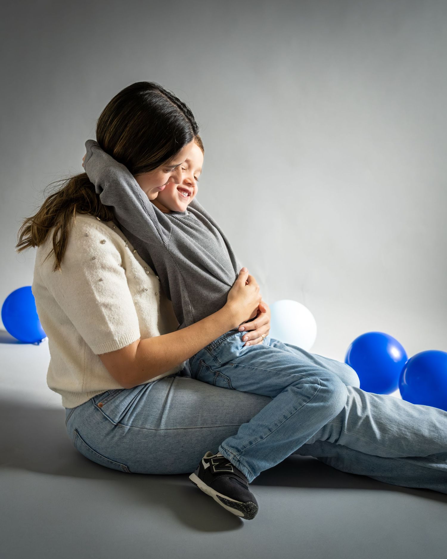 A person in jeans and white shirt lying down with blue balloons in a minimalist studio setting.