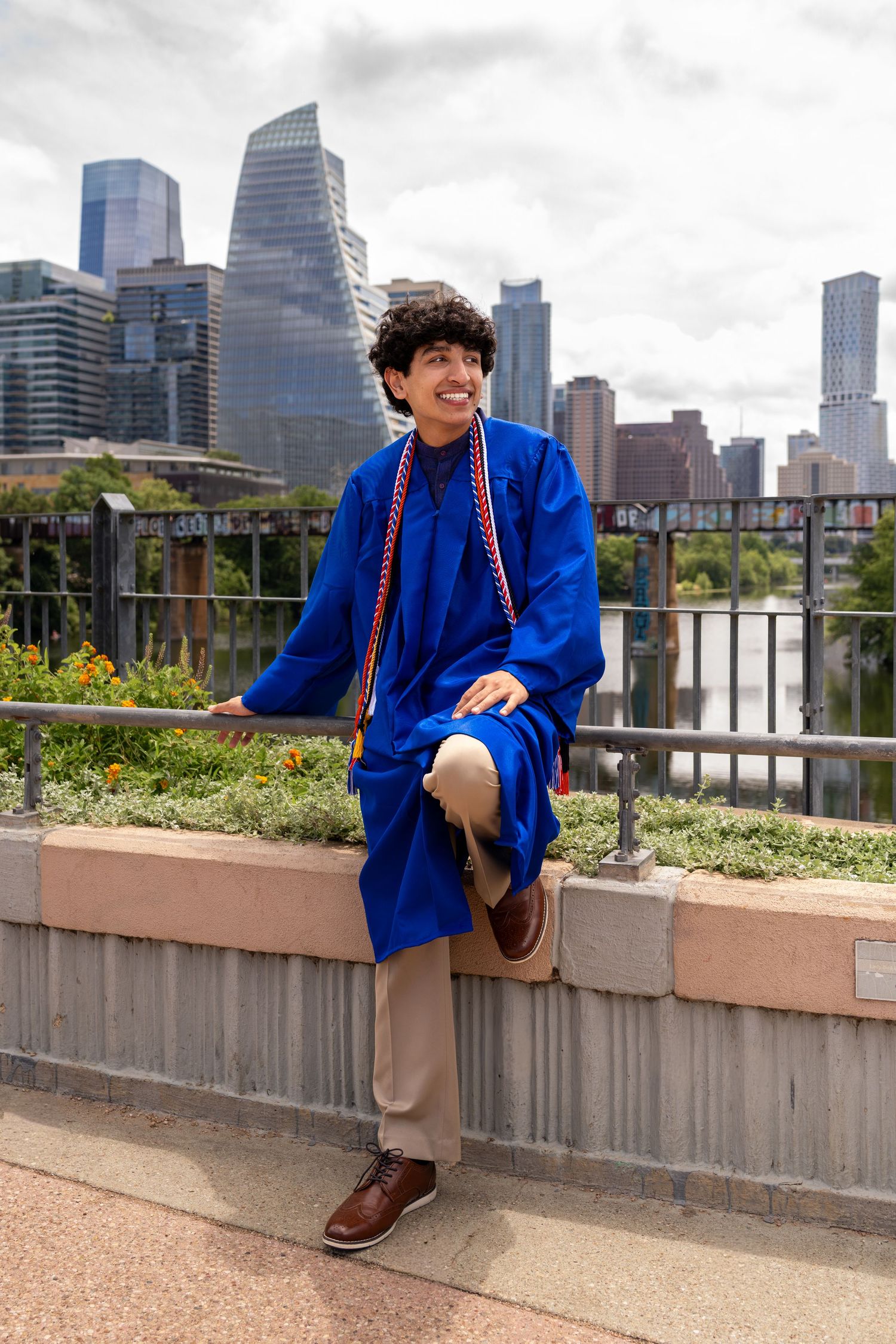 A smiling graduate in a blue cap and gown poses against a city skyline backdrop wearing honor cords.
