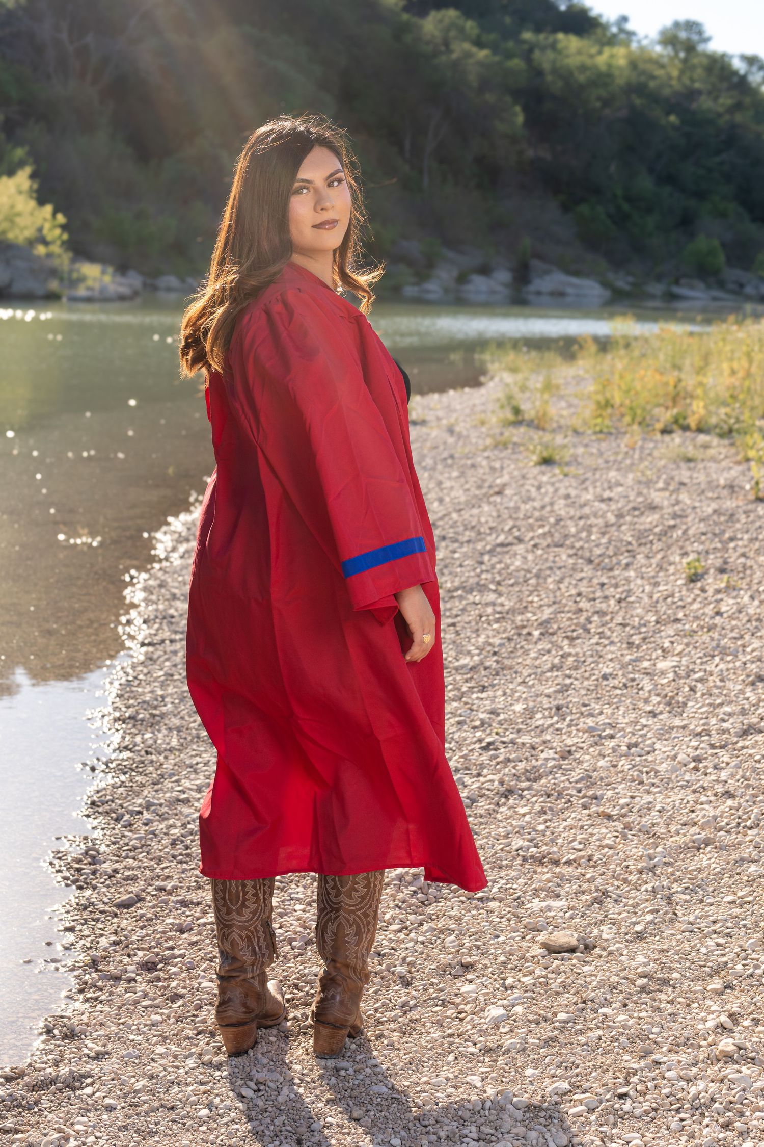 Woman in red midi dress and brown boots standing on rocky riverbank with sunlit hills in background.