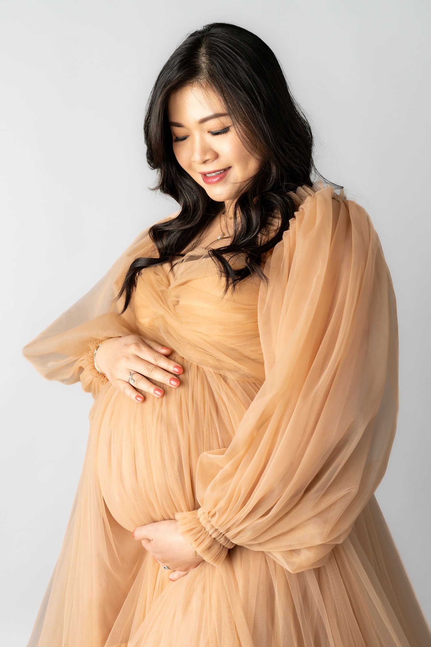 Pregnant woman in elegant golden tulle maternity gown cradling her baby bump, smiling softly against a white background.
