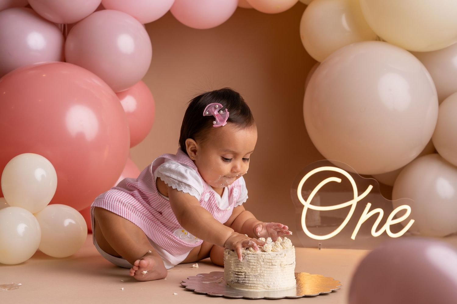 Baby girl digging into white cake at a Life Happens Photography cake smash in Buda, TX, with pink balloon garland and neon sign background.