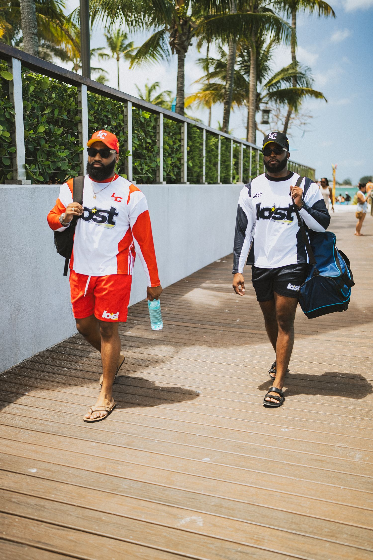 Two men wearing matching 'Lost' brand shirts walk along a sunny boardwalk lined with palm trees.