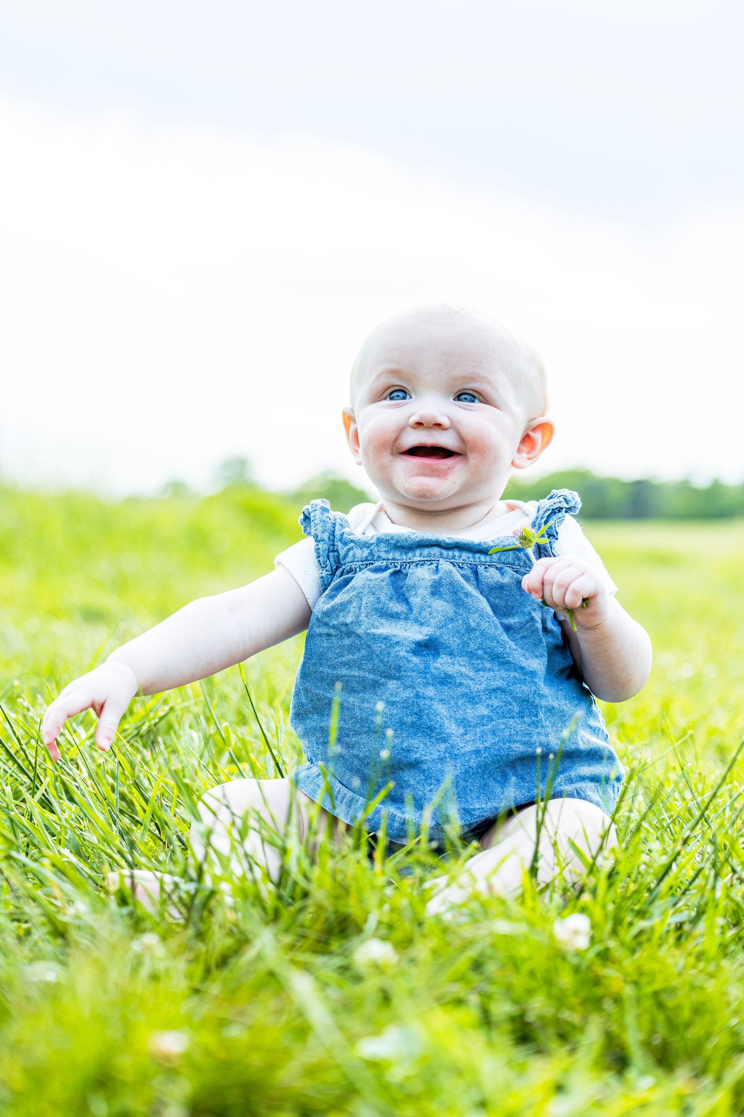 Smiling baby girl with blue eyes sitting in green grass wearing a blue denim dress on a sunny day.
