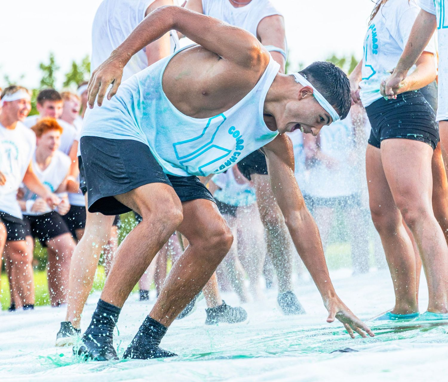 Young man sliding through water at an outdoor color run event surrounded by other participants.