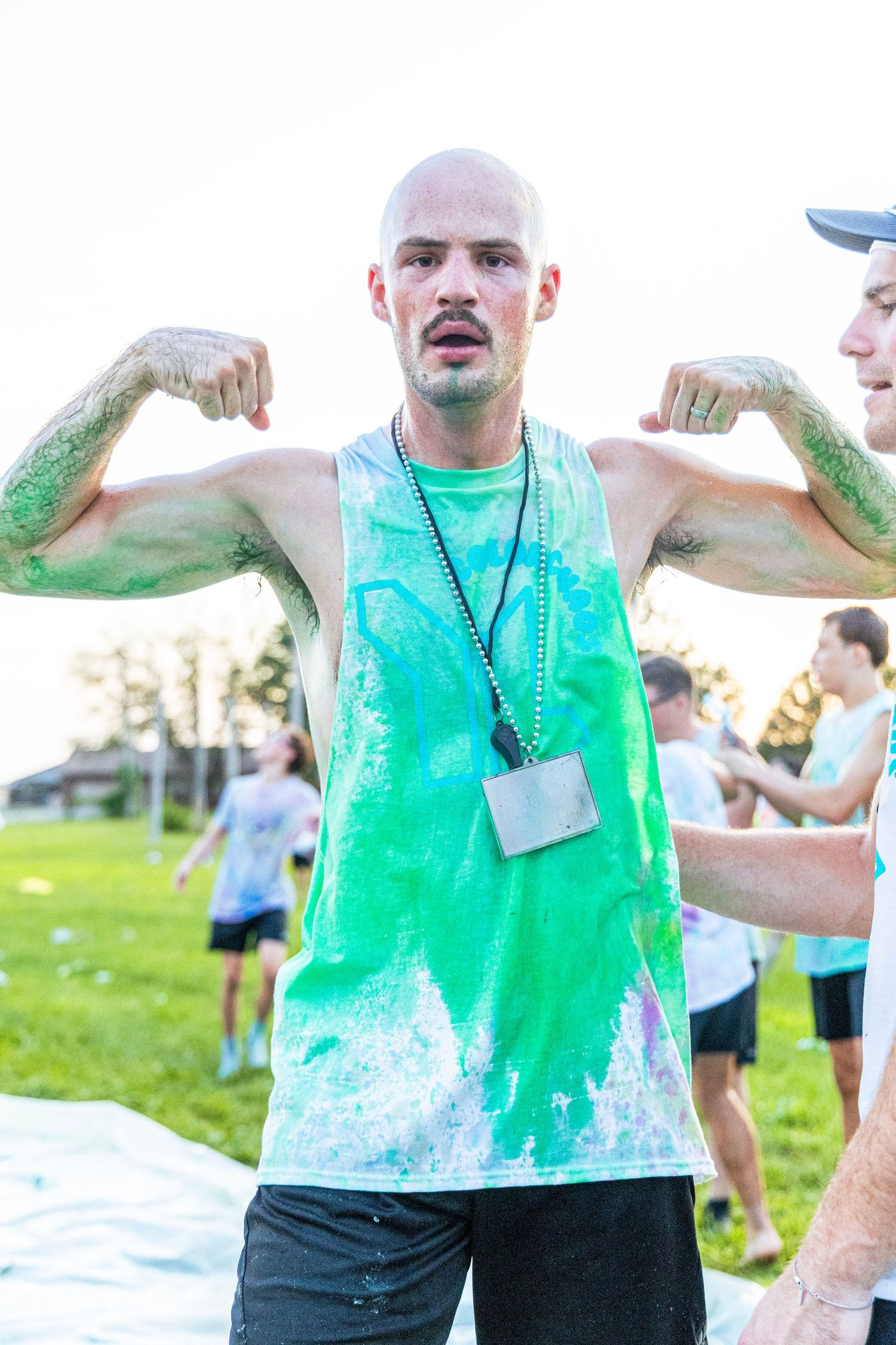 Bald muscular man flexing biceps wearing a green paint-splattered tank top at an outdoor color run event.