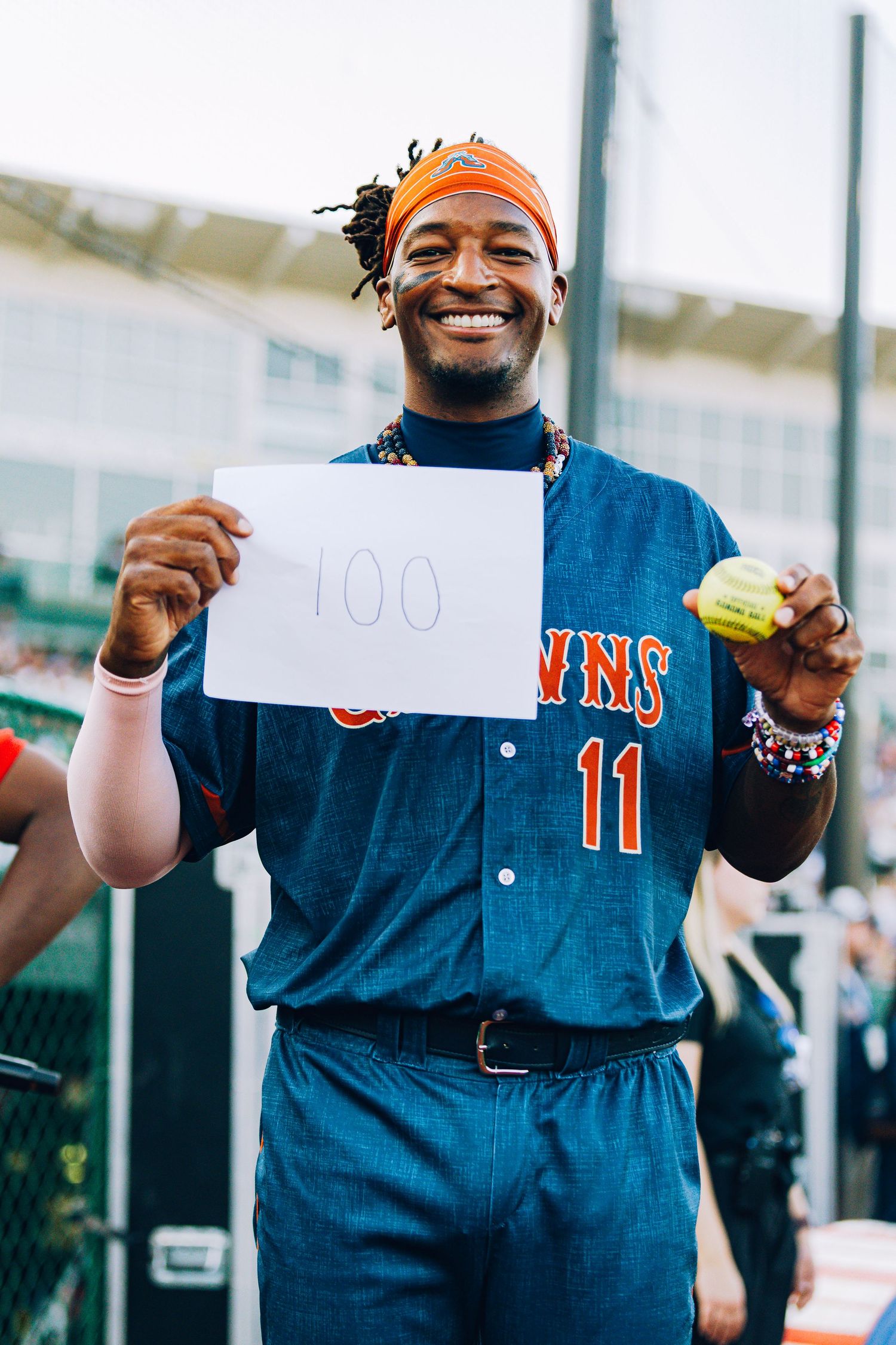A smiling baseball player holds a sign reading 100 and a tennis ball in his jersey.