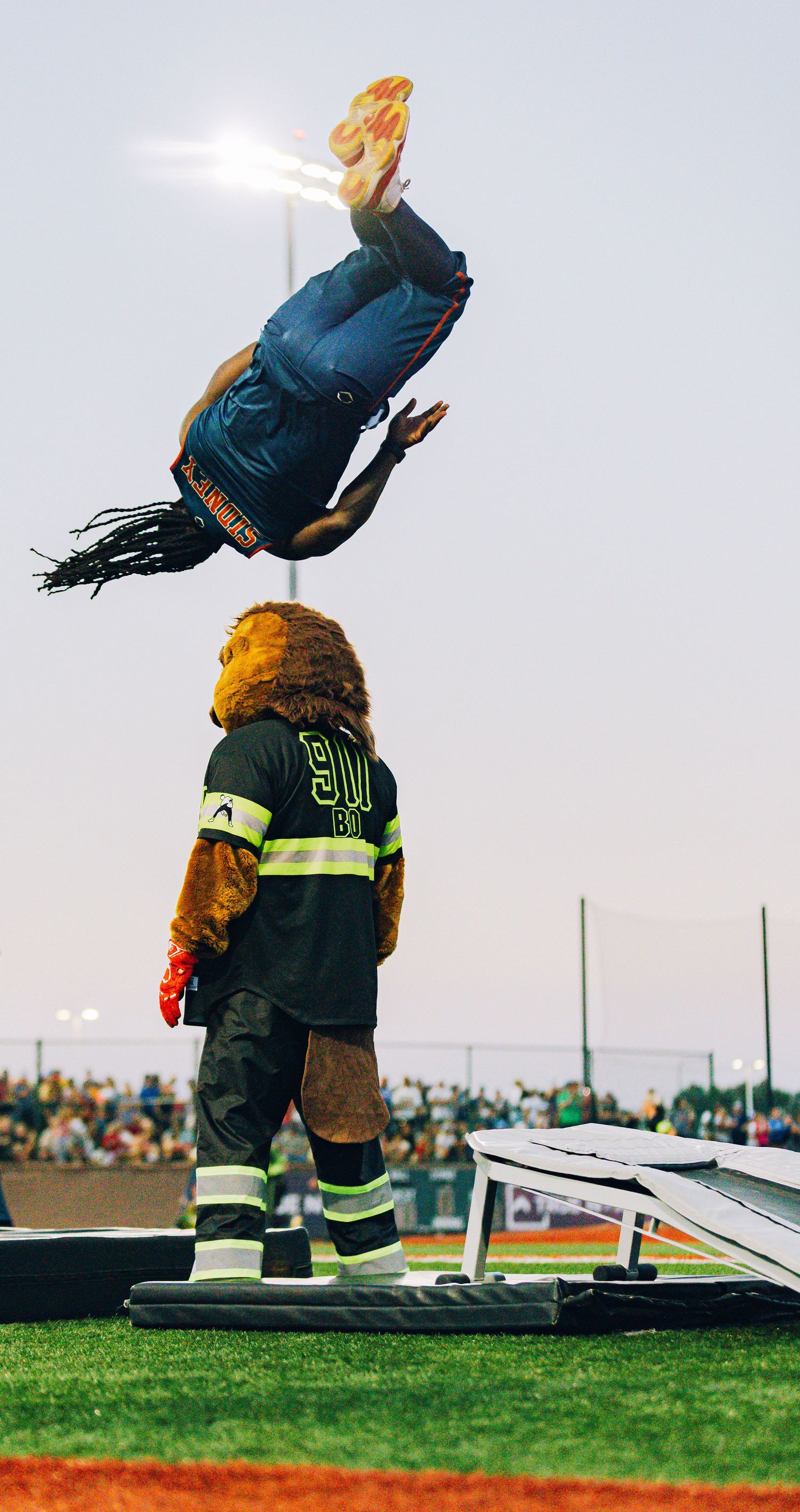 Acrobat performing a backflip over a lion mascot in firefighter gear at a stadium event.