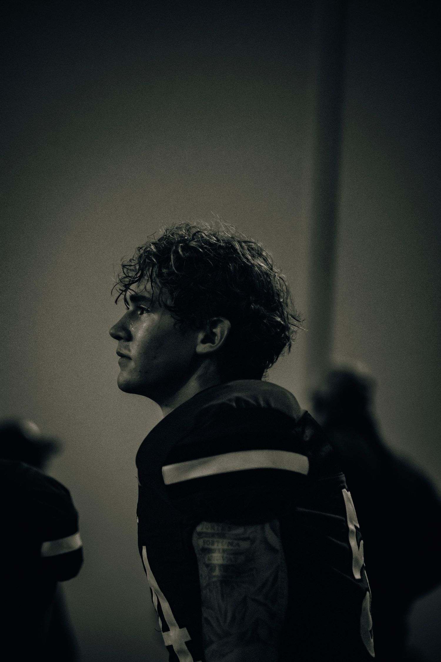 A moody black and white side profile of a curly-haired football player in uniform.