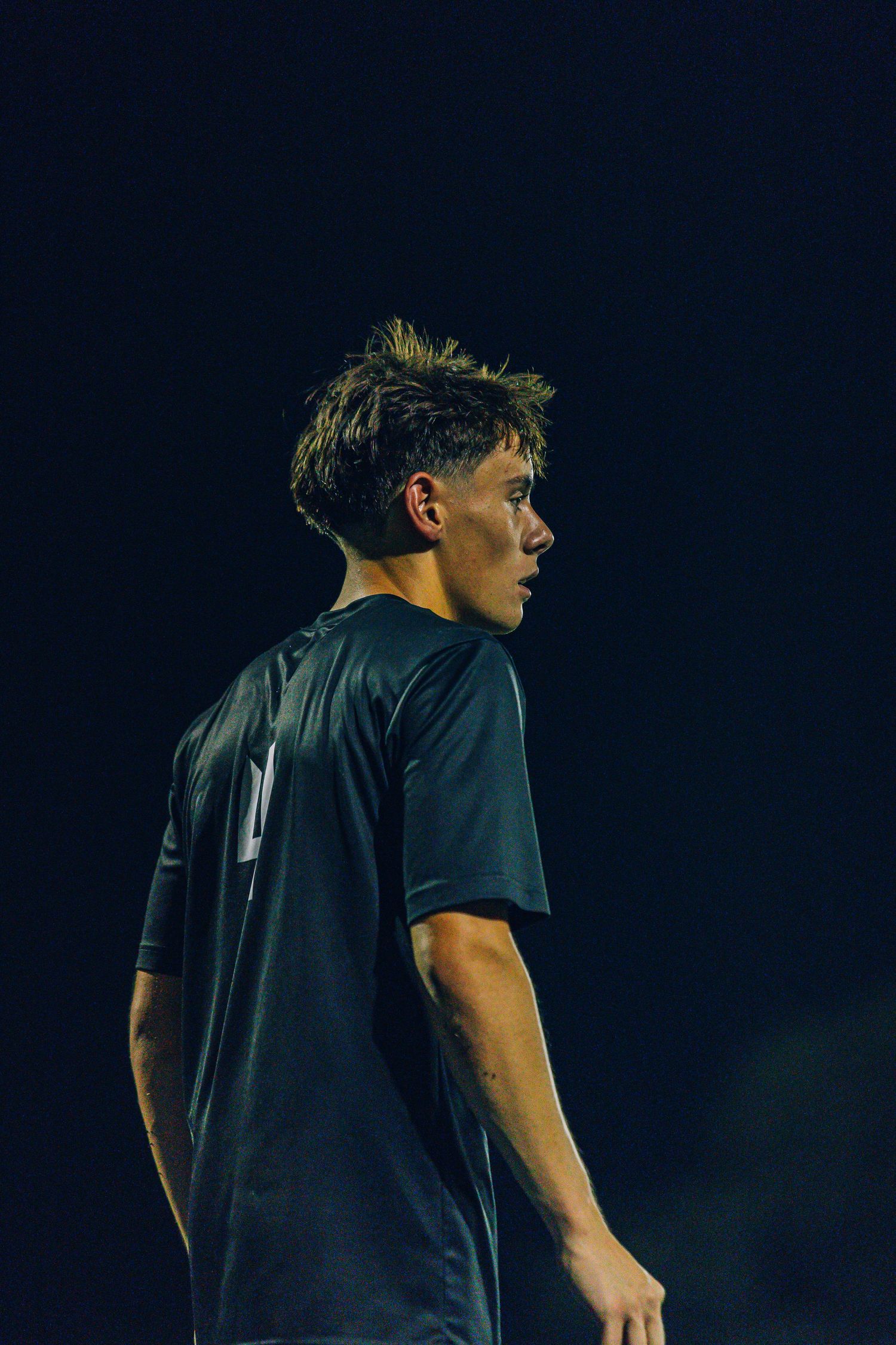 Young male soccer player in black jersey looks over his shoulder under night game lights.