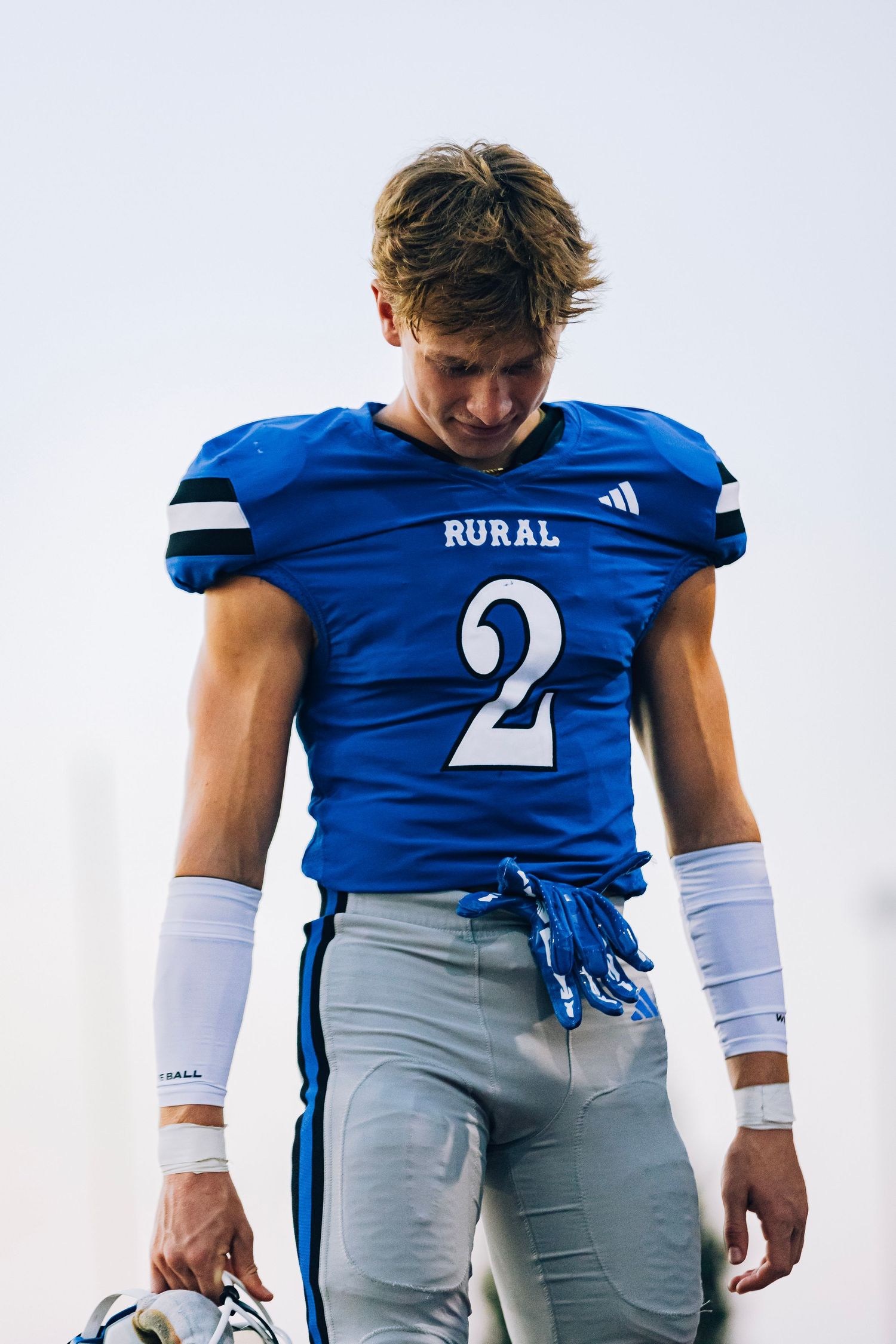 A high school football player wearing a blue Rural jersey number 2 with white wristbands looks down on the field.