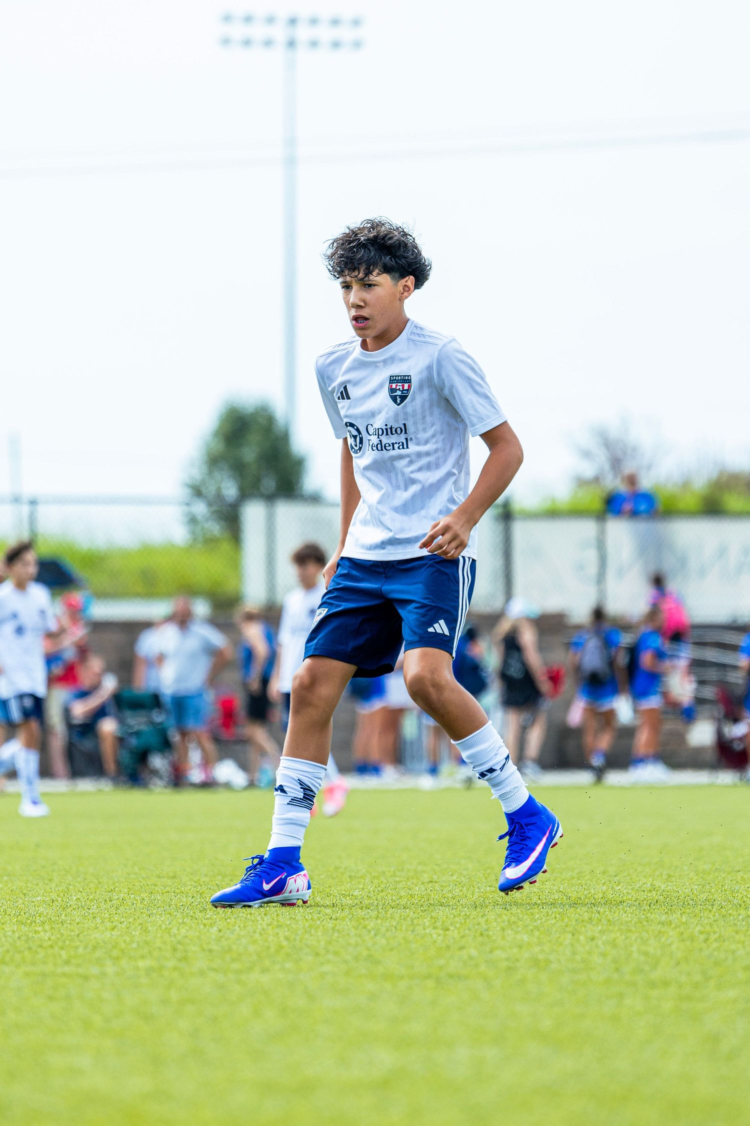 Young male soccer player in white Sporting KC jersey and blue shorts dribbling on a green field.