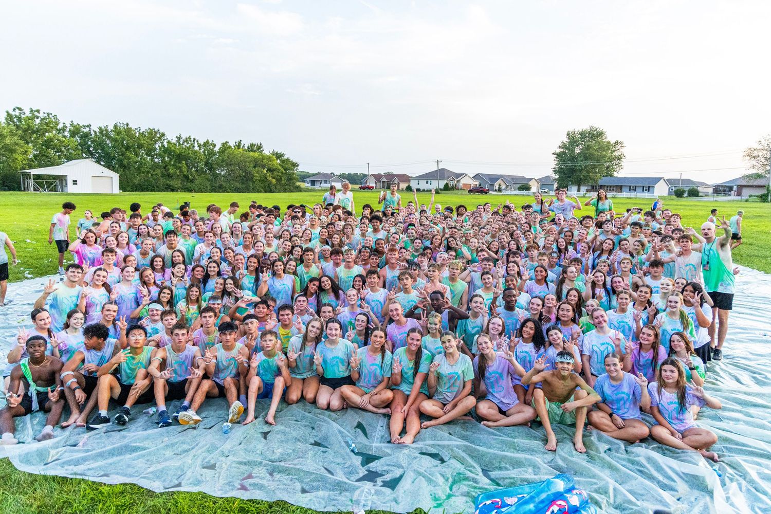 Large group of students in matching teal tie-dye shirts posing together outdoors on a sunny day.