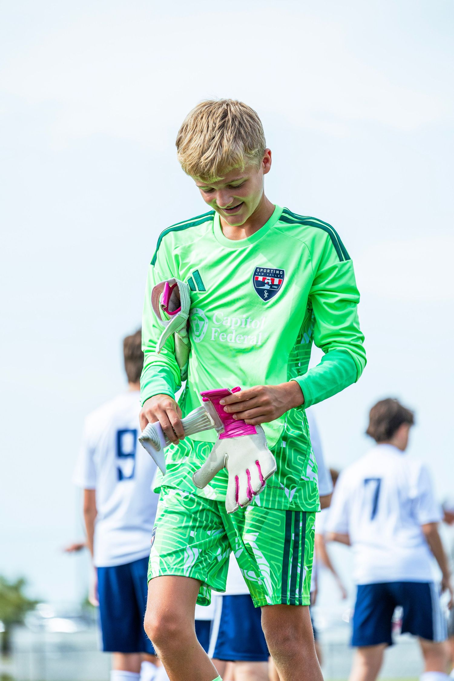 Young blonde goalkeeper in neon green Sporting KC uniform holding gloves on a sunny soccer field.