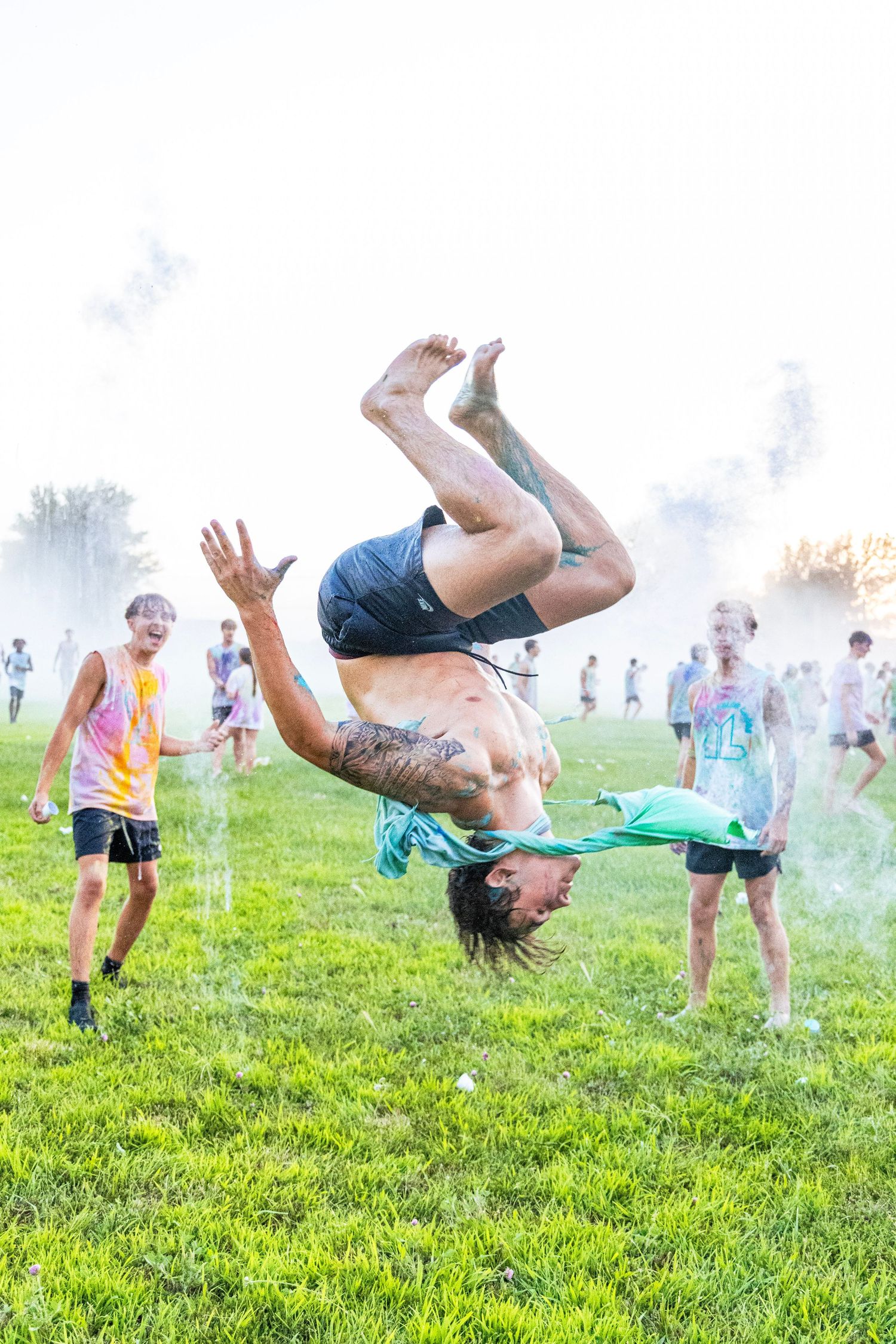 Tattooed man performing a backflip at an outdoor color powder festival surrounded by cheering kids.