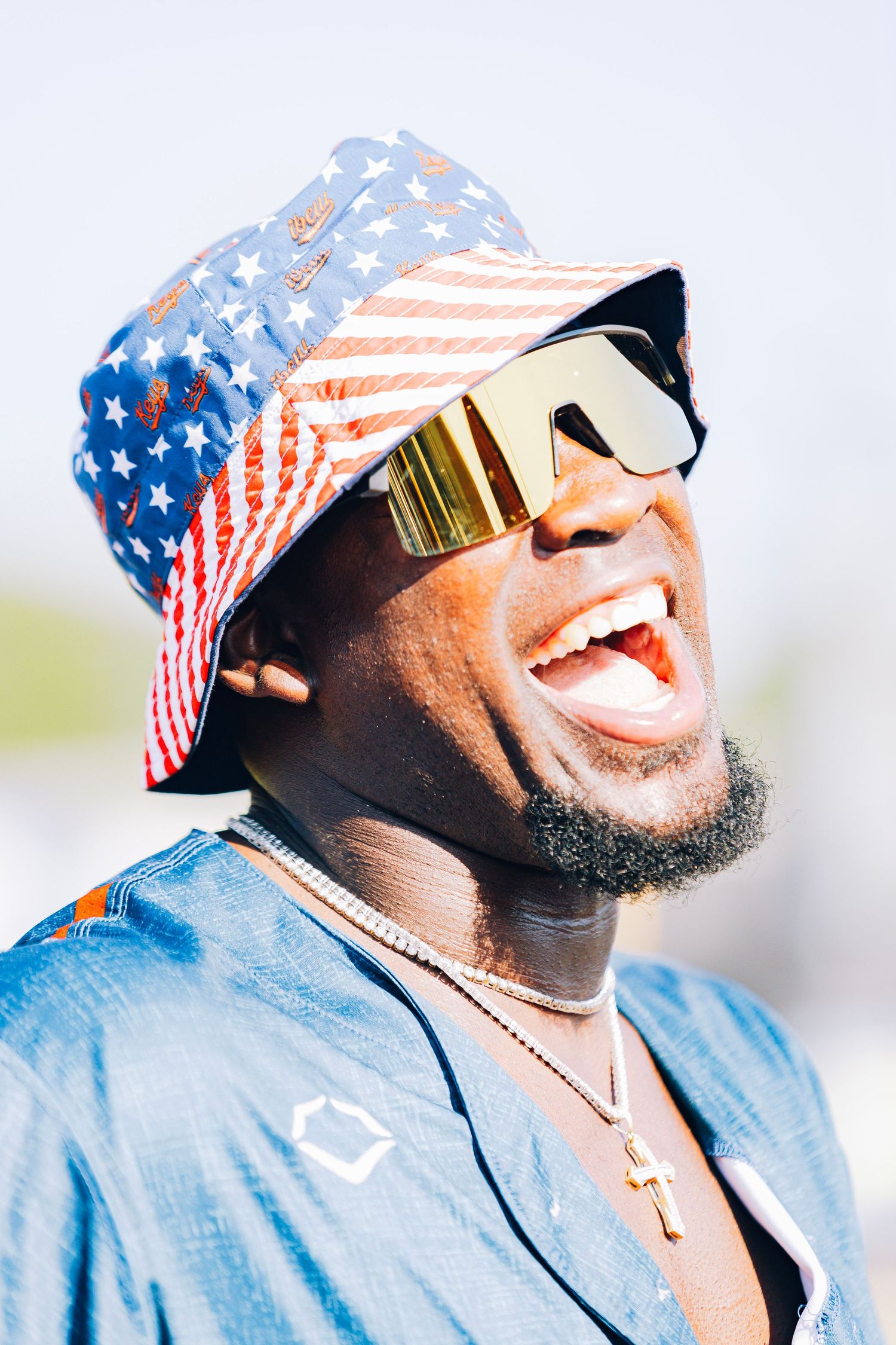 Laughing Black man wearing an American flag bucket hat and gold mirrored sunglasses outdoors.