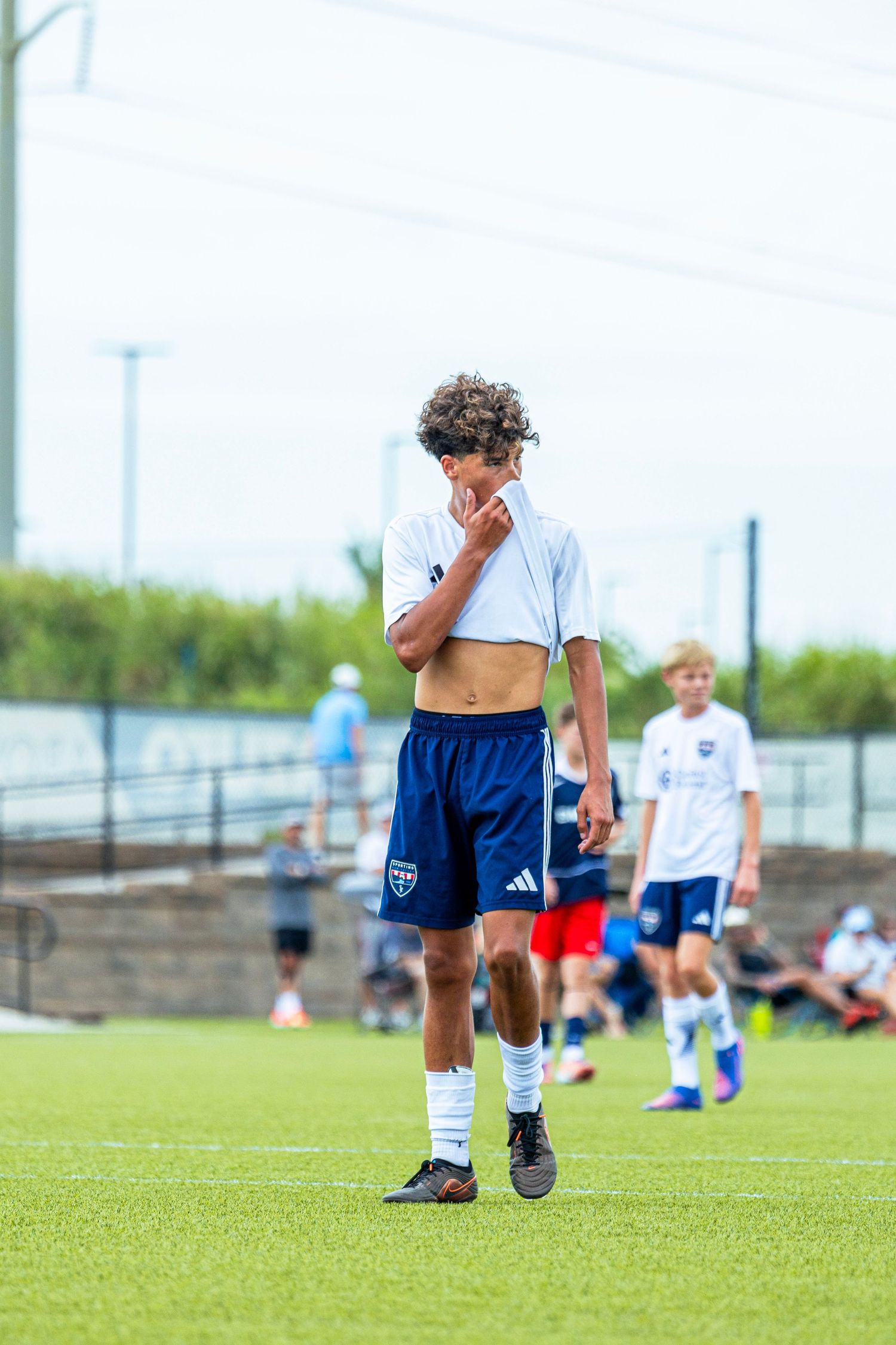 A curly-haired teen soccer player lifting his shirt to wipe his face during a match.
