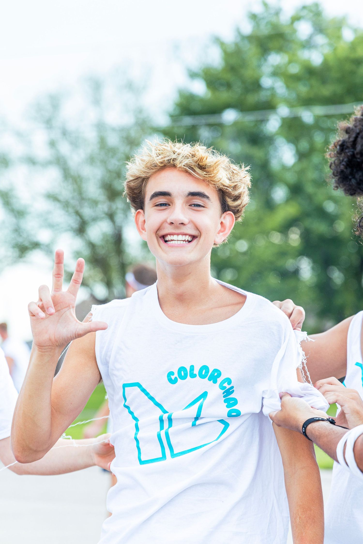 Smiling teenage boy in a white Color Chaos t-shirt giving a peace sign at an outdoor event.