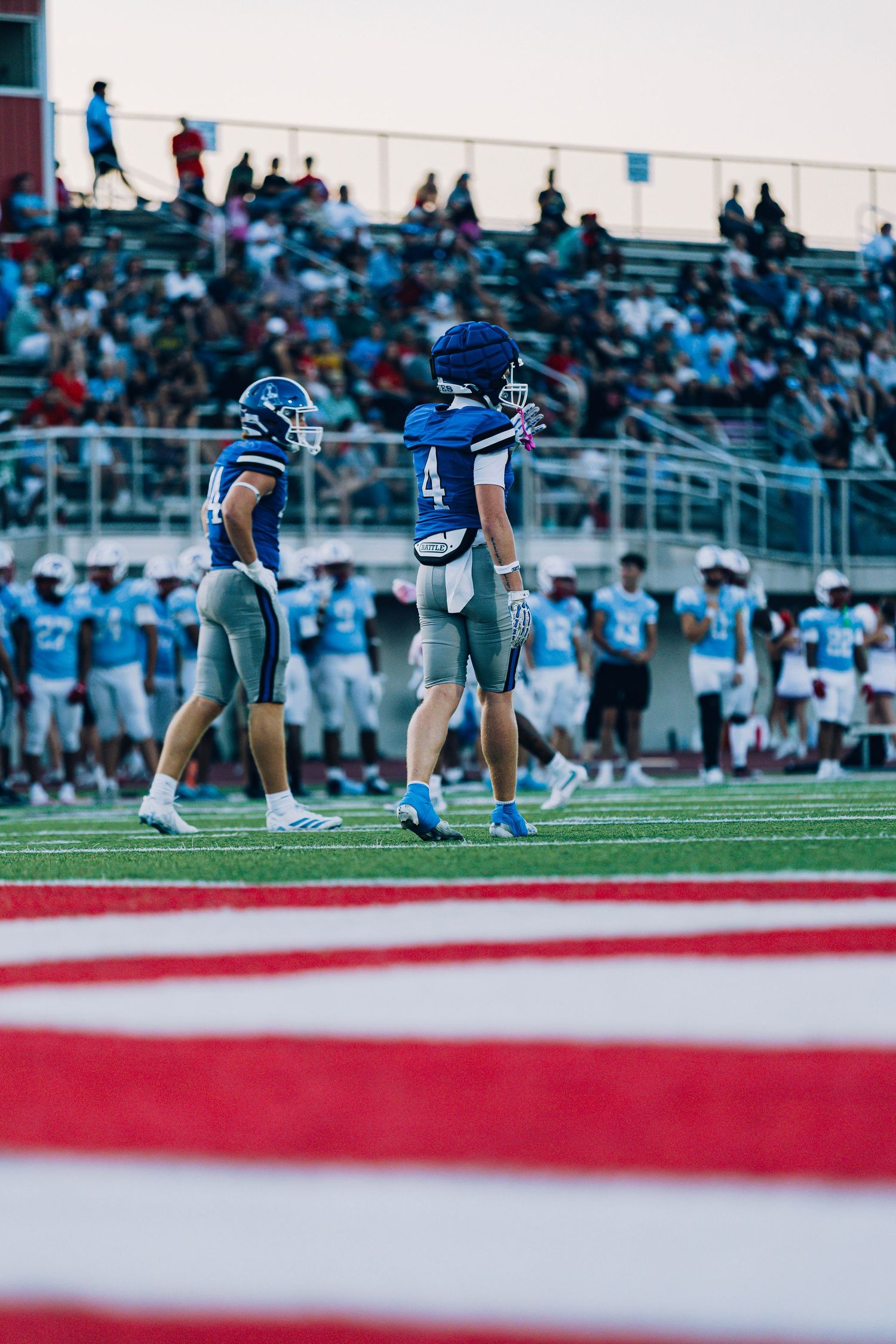 Two football players in blue uniforms on field with packed stadium bleachers in background.
