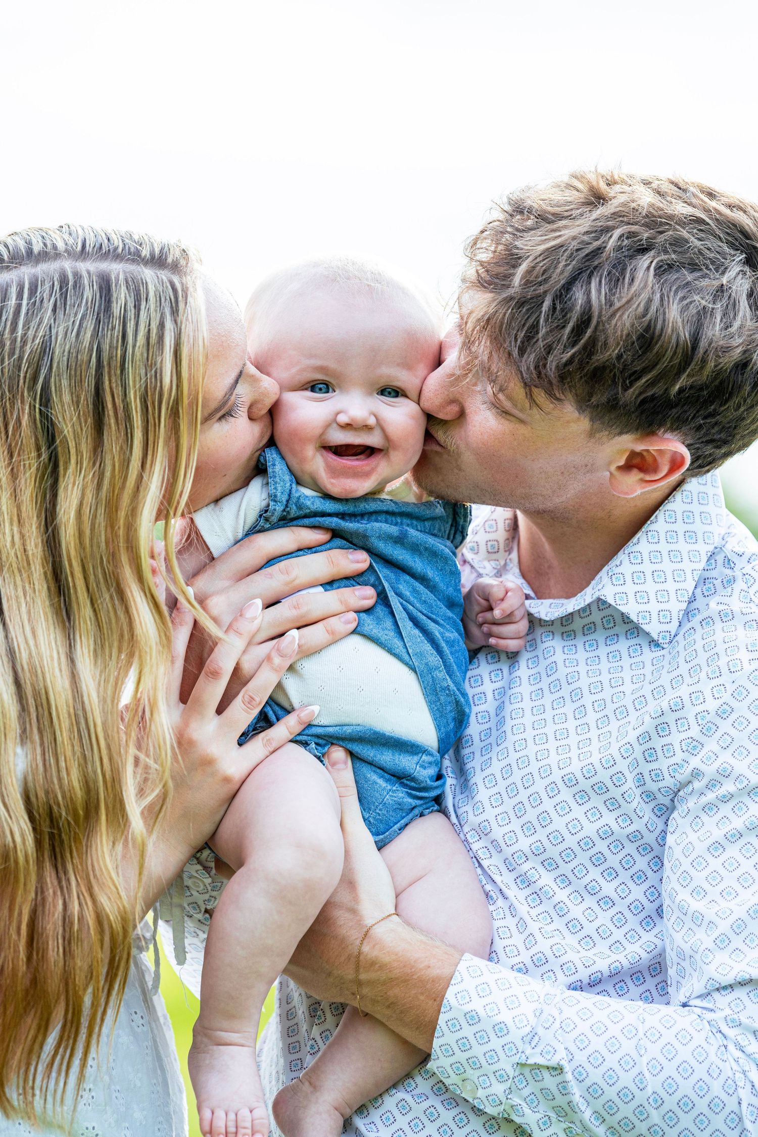 Happy baby smiling while parents kiss both cheeks in a sweet outdoor family portrait.