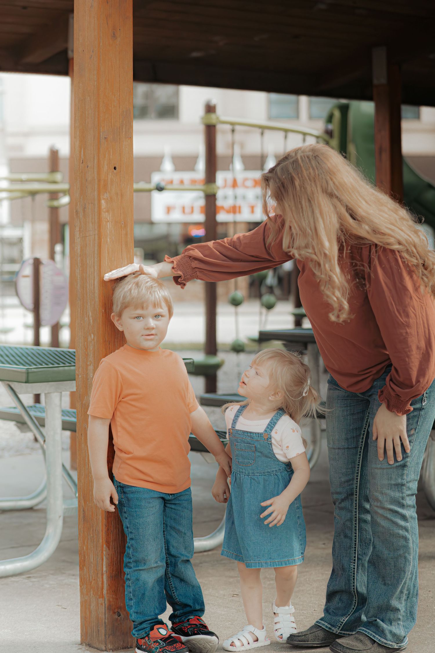 A woman measures a young boy's height against a wooden post while a toddler girl watches at an outdoor playground.