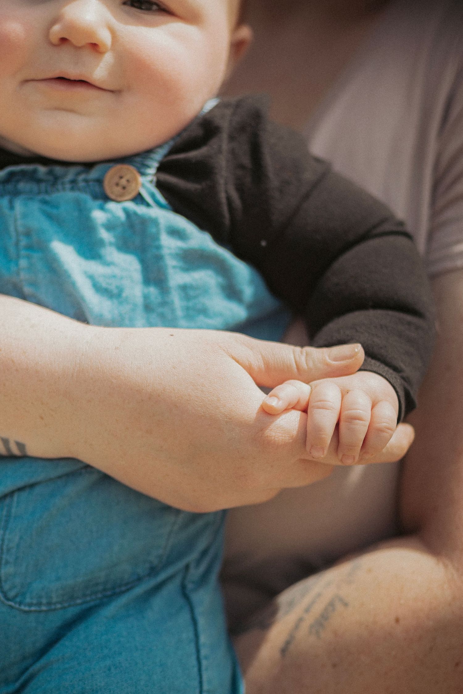 A smiling baby in denim overalls holds an adult's hand, showcasing a tender moment between parent and child.
