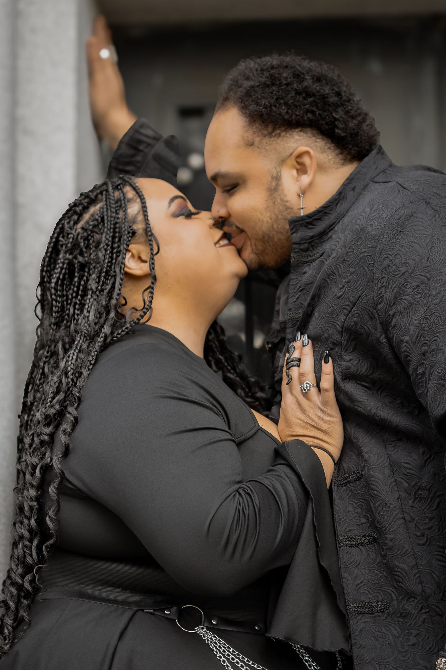 A couple dressed in black sharing a romantic kiss, with the woman wearing braids and rings.