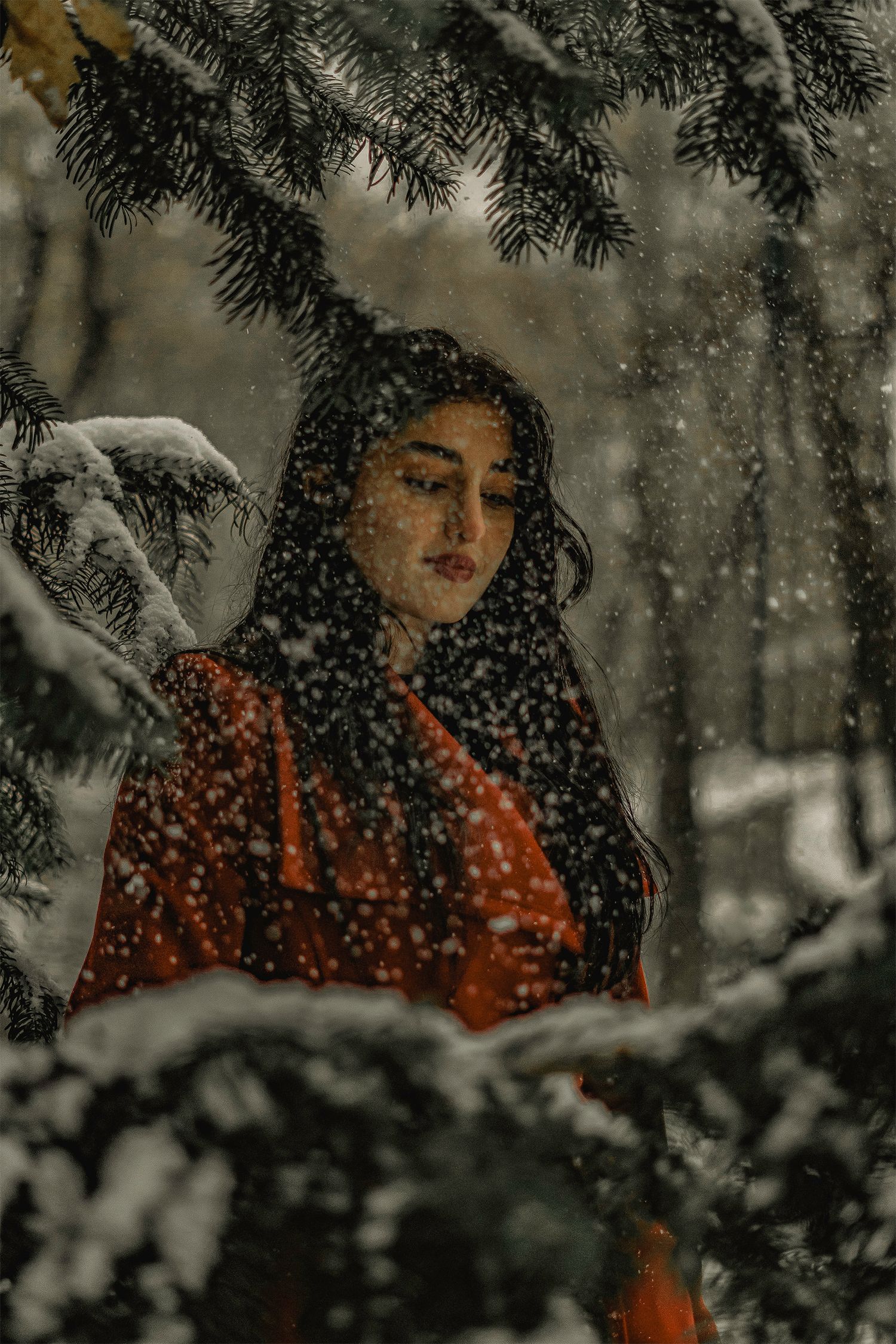 Woman in red coat standing in snowy forest with falling snow and pine trees.